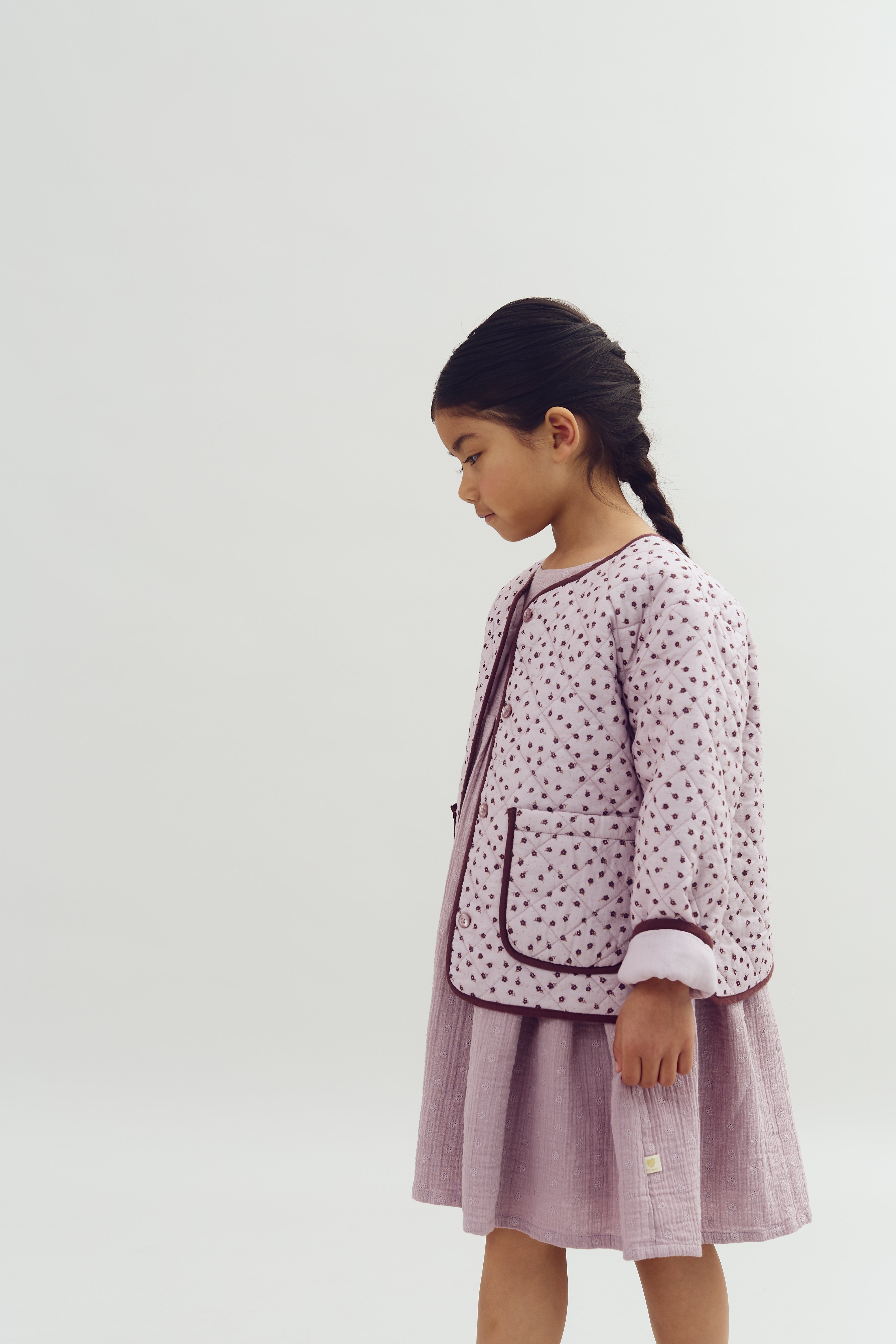 A young girl, wearing a patterned jacket and dress, stands in profile with hands at her sides against a plain, light background.