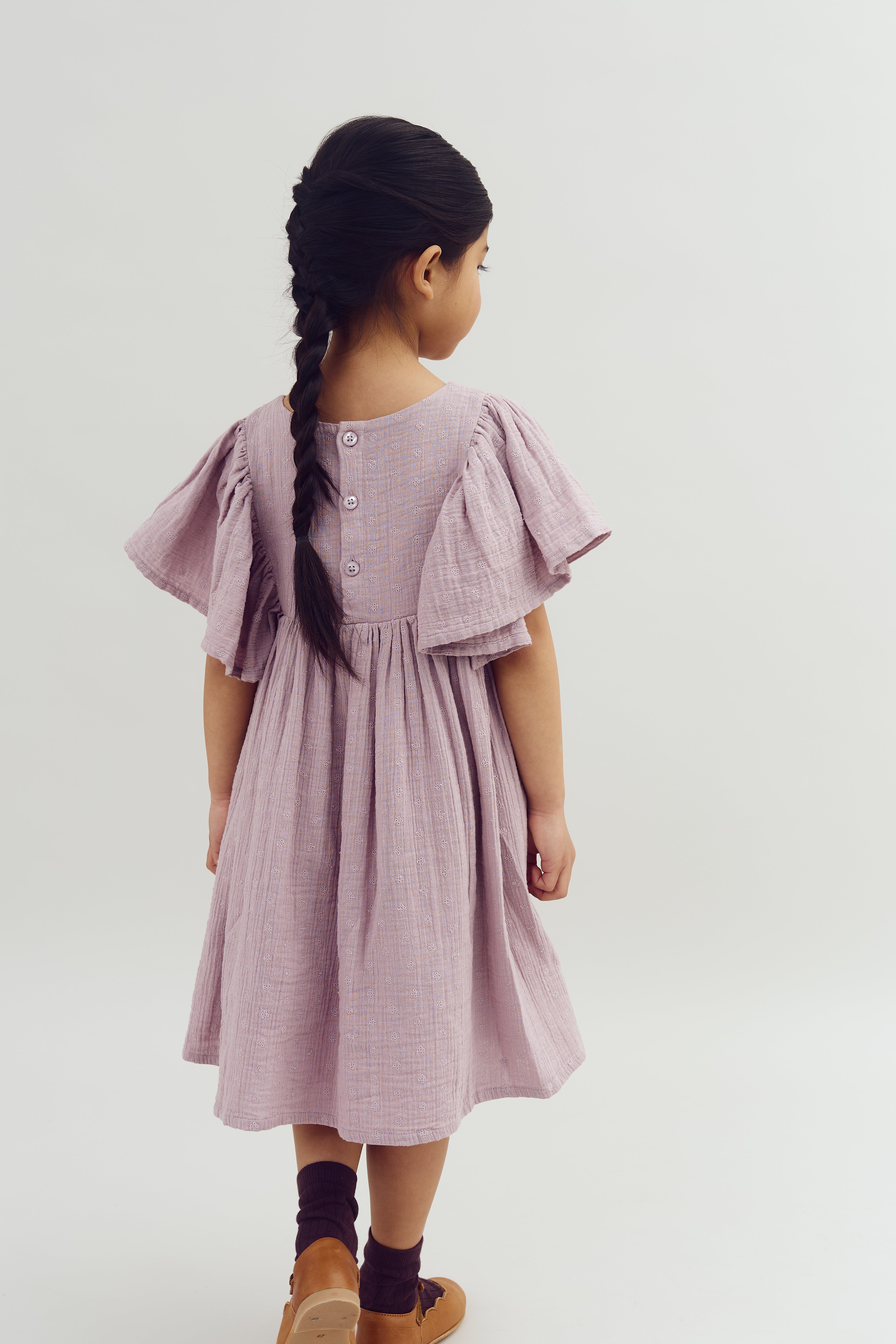 A young girl with a braided hairstyle stands facing away, wearing a lilac dress with billowy sleeves and buttons on the back, in a plain, neutral background.