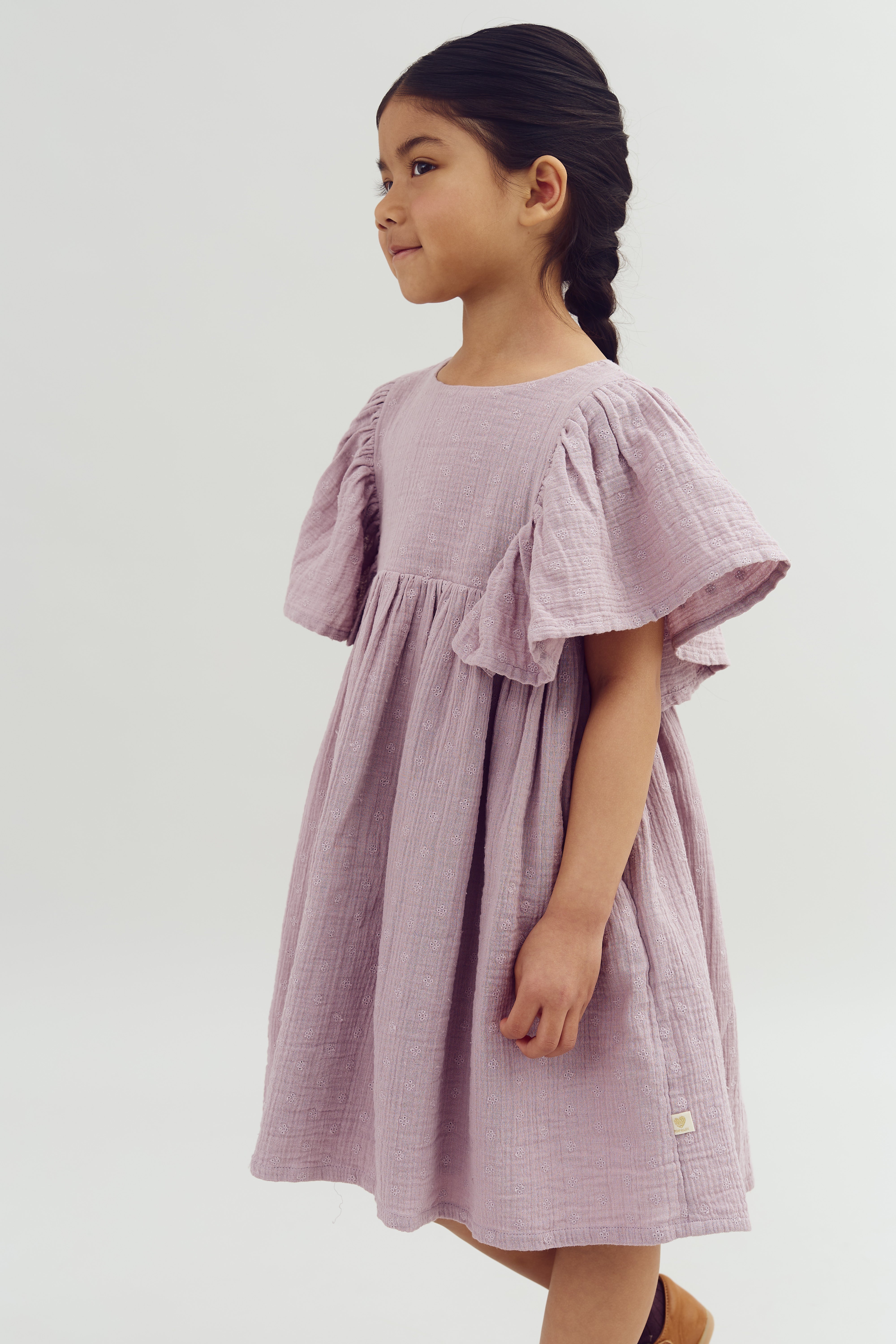 A child in a textured, lilac dress with flutter sleeves stands in profile, featuring braided hair. The background is a plain, light gray, creating a soft, minimalistic setting.