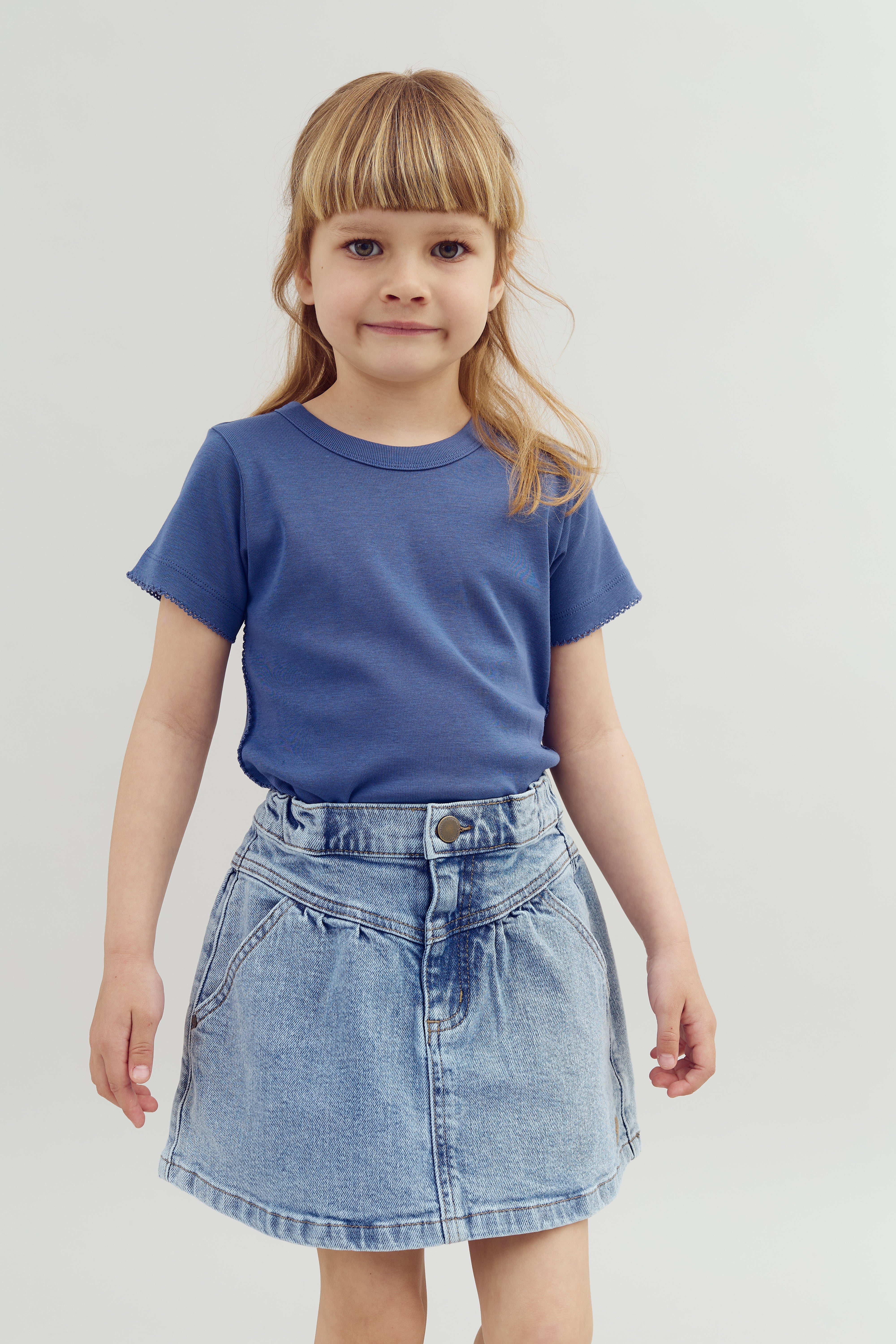 Barn står stille iført en blå t-shirt og en nederdel af denim. Baggrunden er neutral og lys, hvilket fokuserer opmærksomheden på barnet.