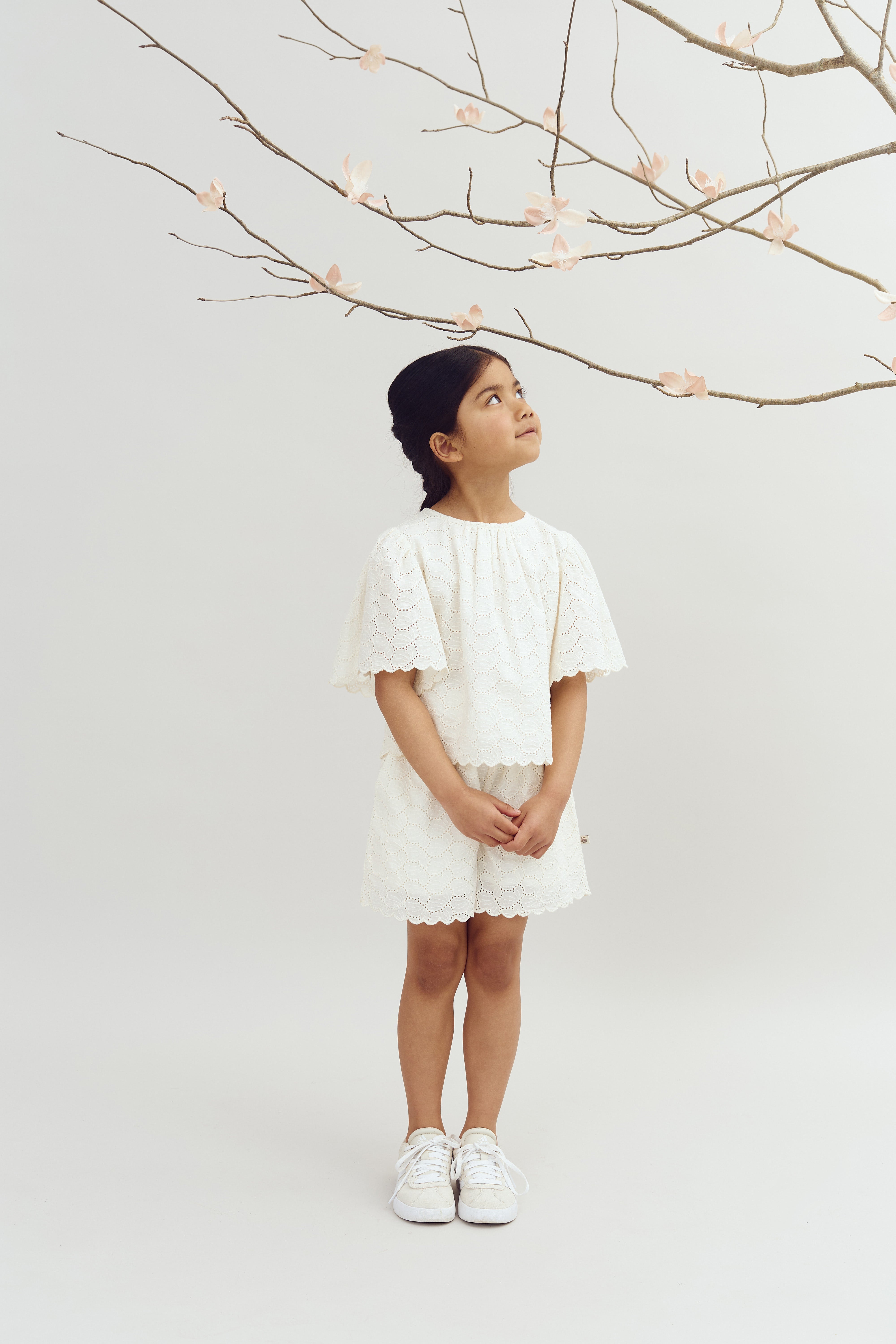 A girl in a white lace outfit is looking up at pink blossoms on a bare branch, set against a plain, light-colored background.