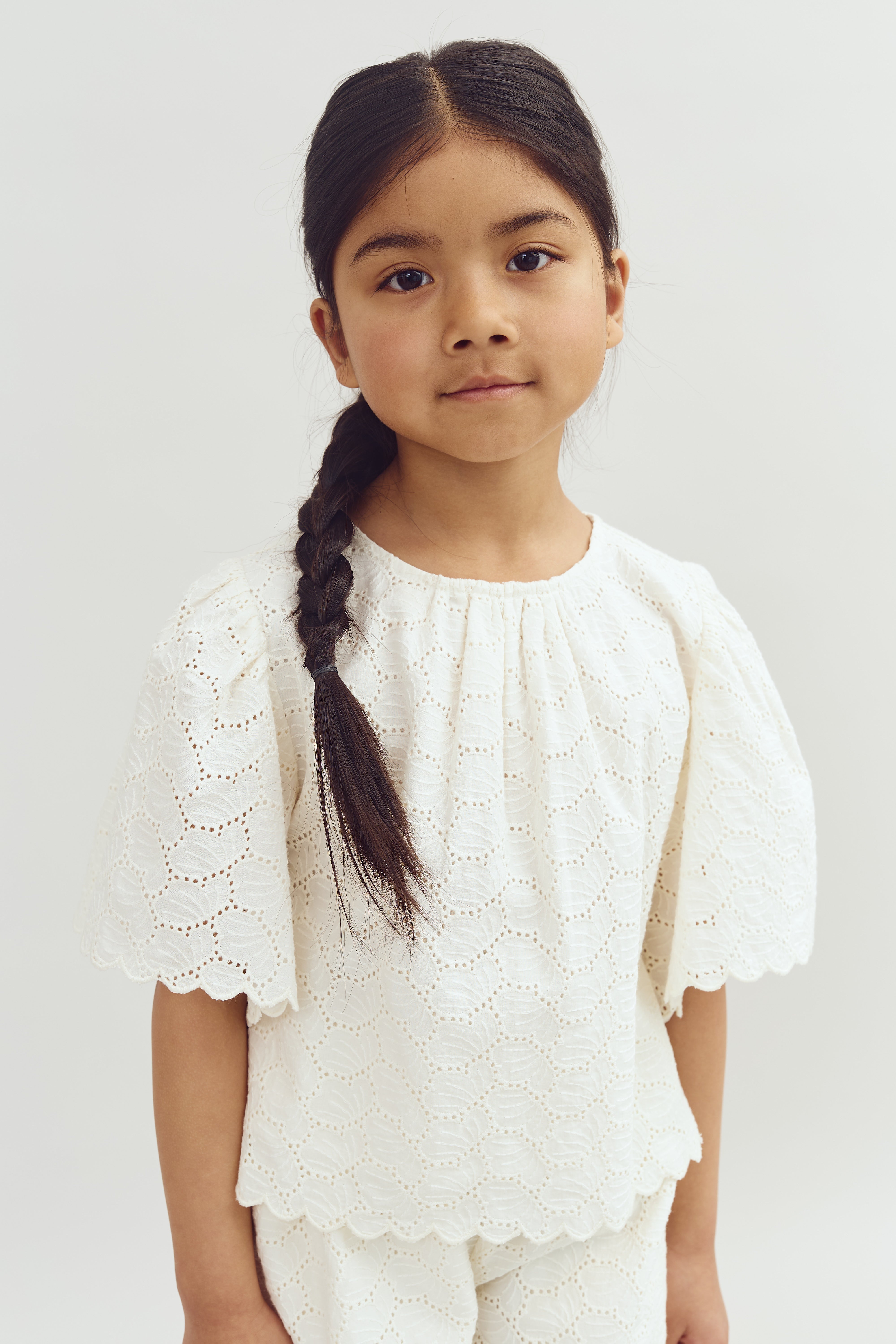 Child stands calmly, wearing a white lace outfit with puffed sleeves and a braid, set against a plain, light background.