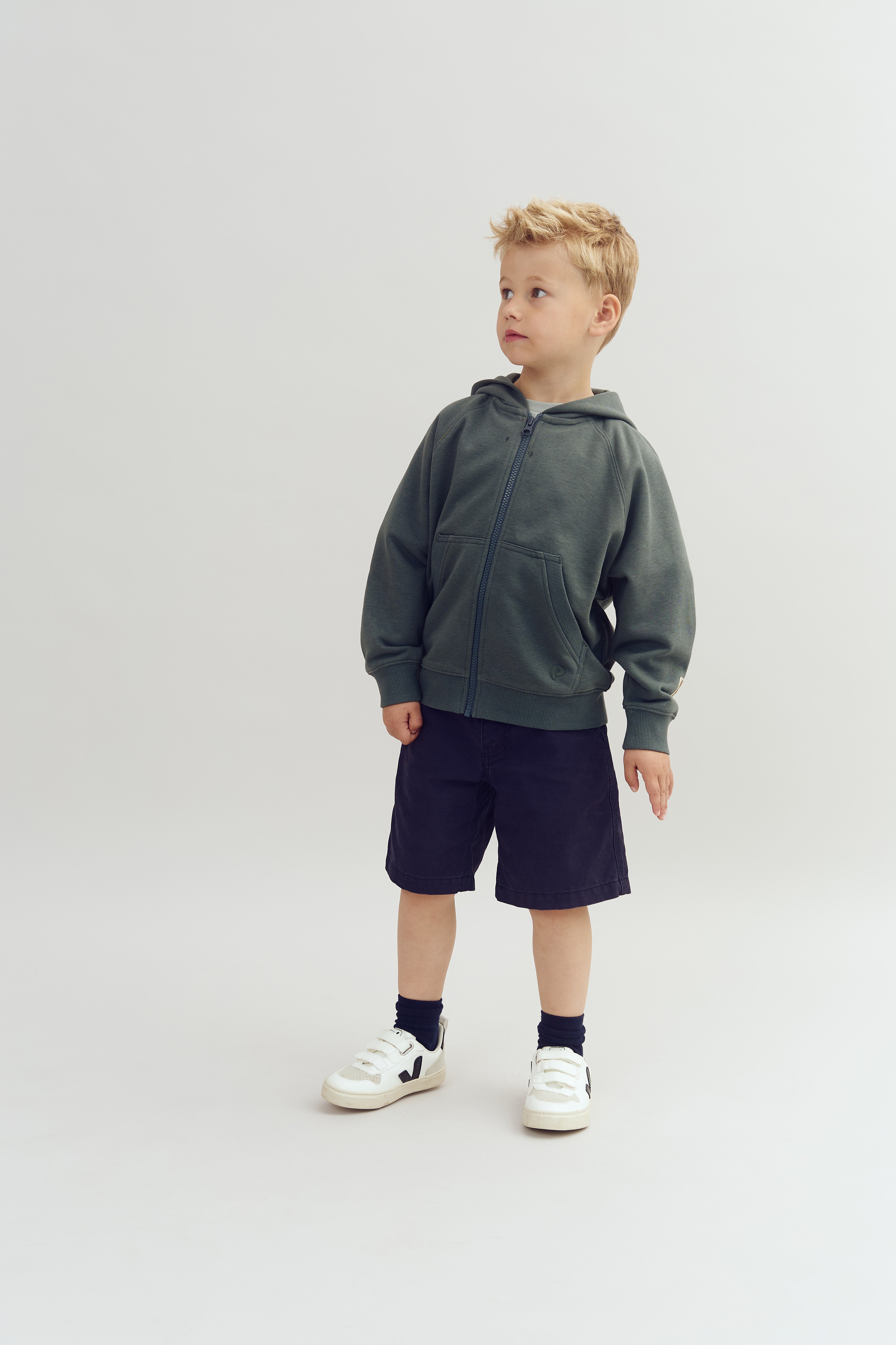 A young child, wearing a dark hoodie and shorts, stands looking to the side. They are in a studio environment with a plain light-colored background.