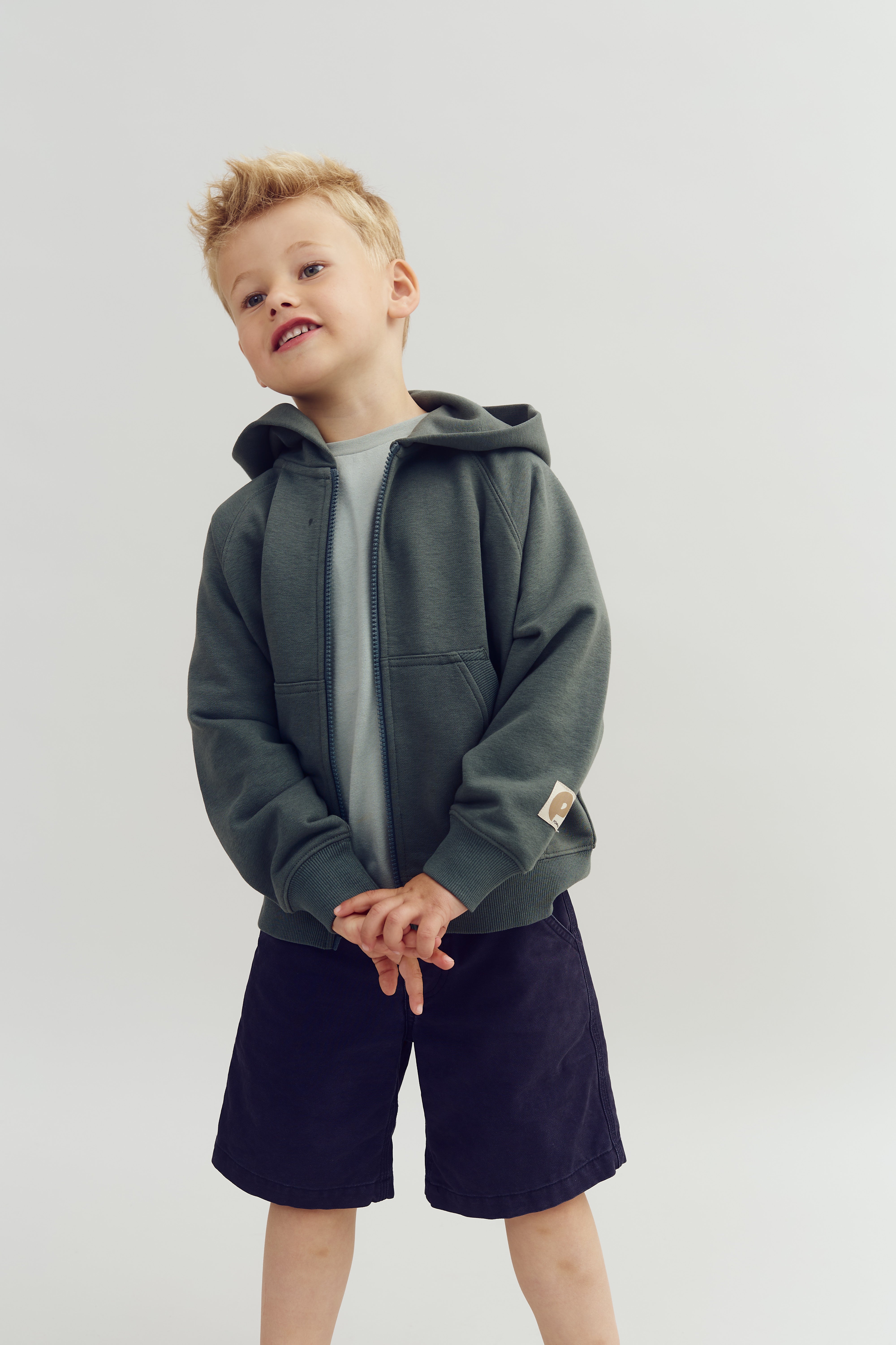 A young child wearing a gray hooded sweatshirt and dark shorts stands with hands clasped, slightly smiling, against a neutral background.