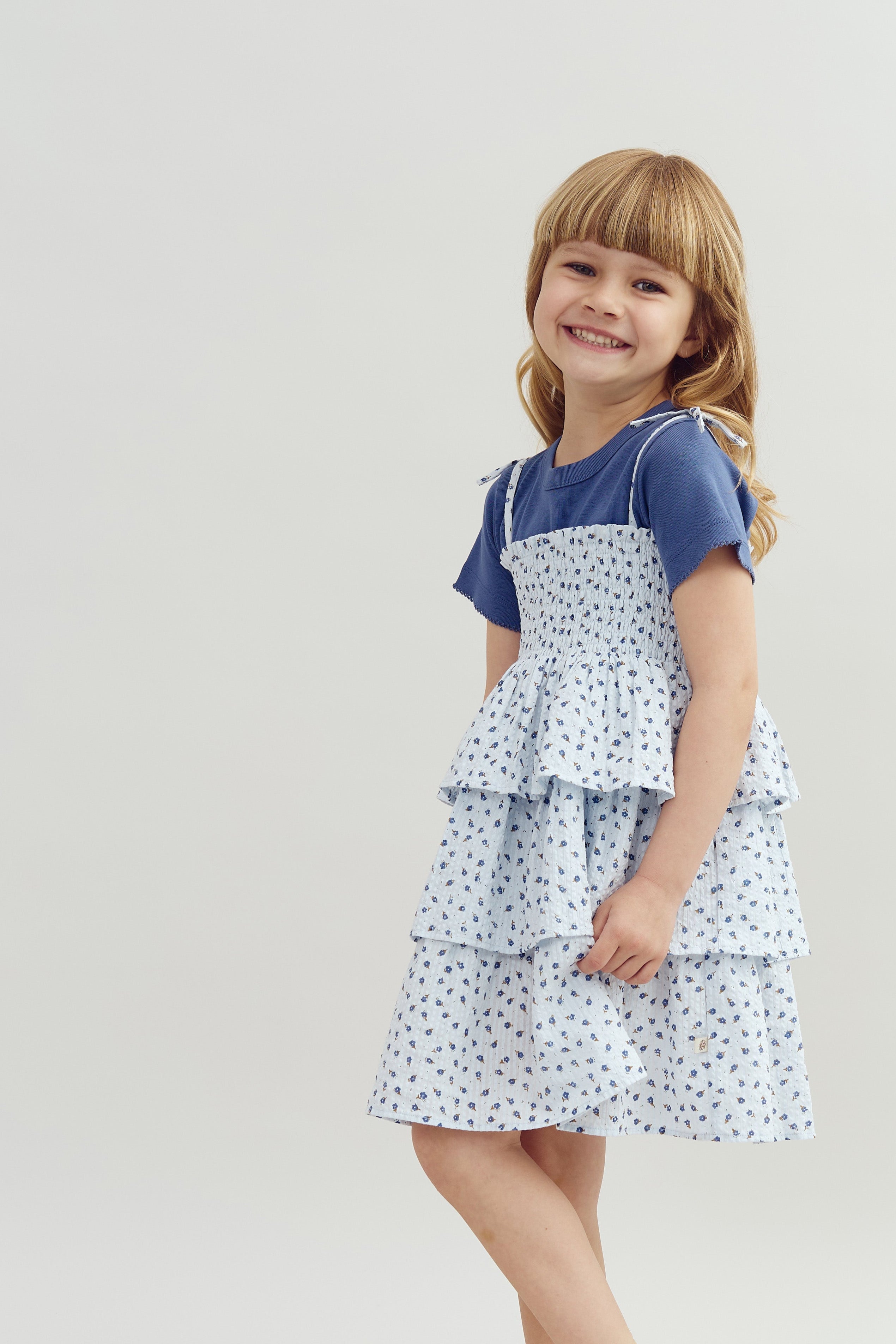 A young girl smiles gently, wearing a blue t-shirt and a tiered floral dress against a plain, light gray background.