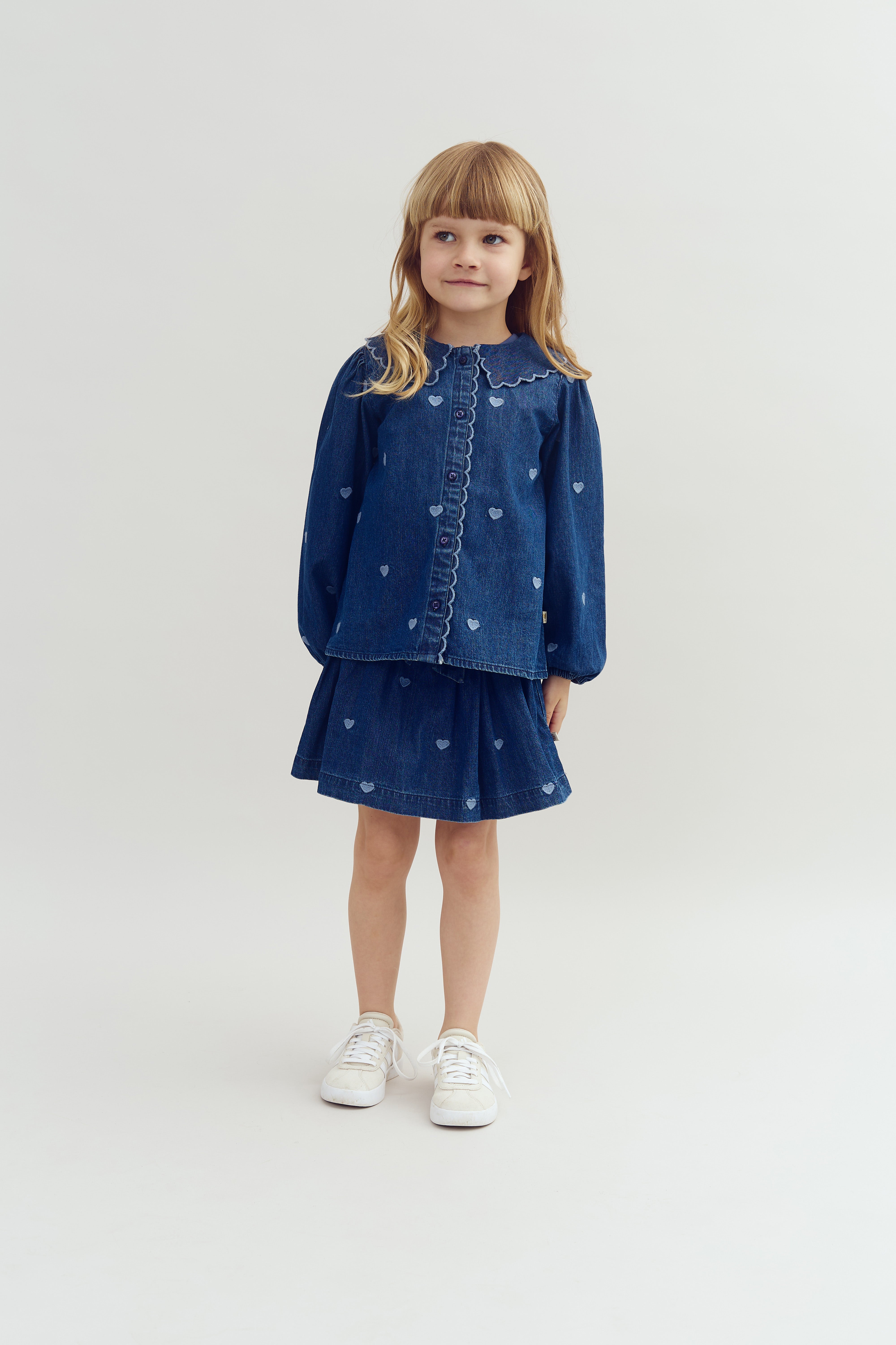 En pige iført en denimkjole med hjerteprint står stille på en lys baggrund, iført hvide sneakers.