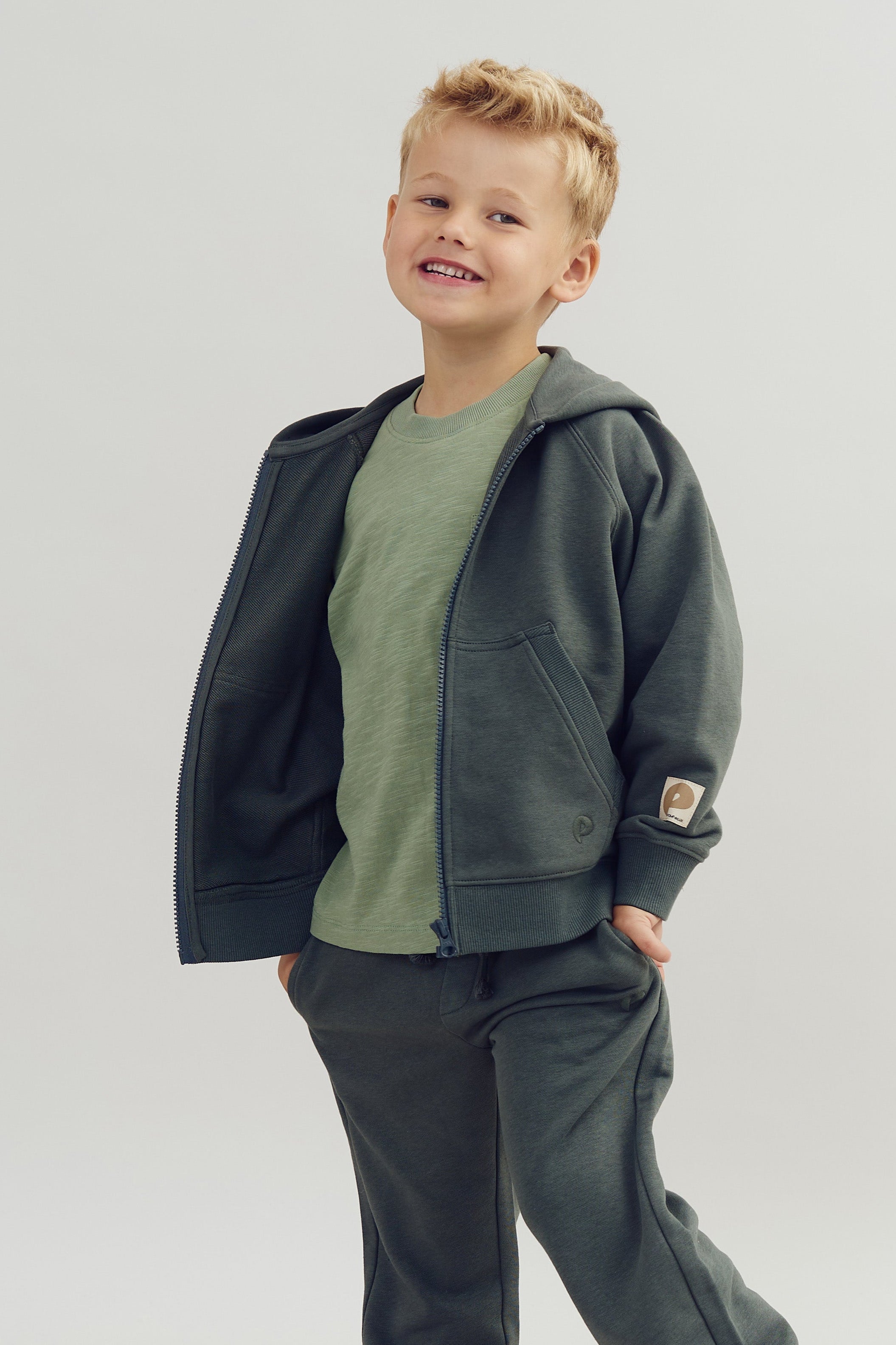 A smiling child wearing a dark gray hoodie and pants, and a light green shirt, stands with hands in pockets against a plain white background.