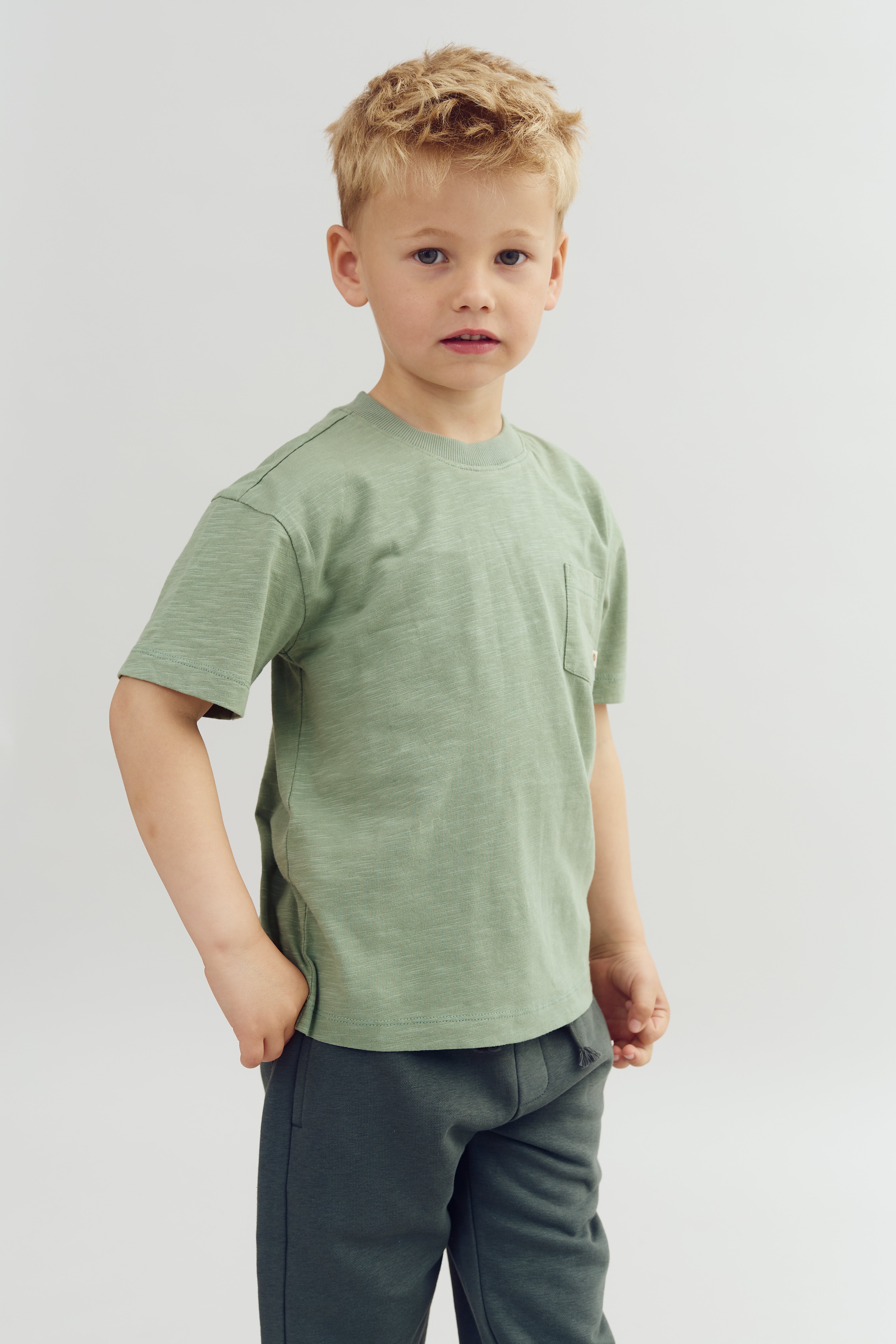 A young child stands with a neutral expression, wearing a light green T-shirt and dark pants. The background is plain and light-colored, emphasizing the child's attire and features.