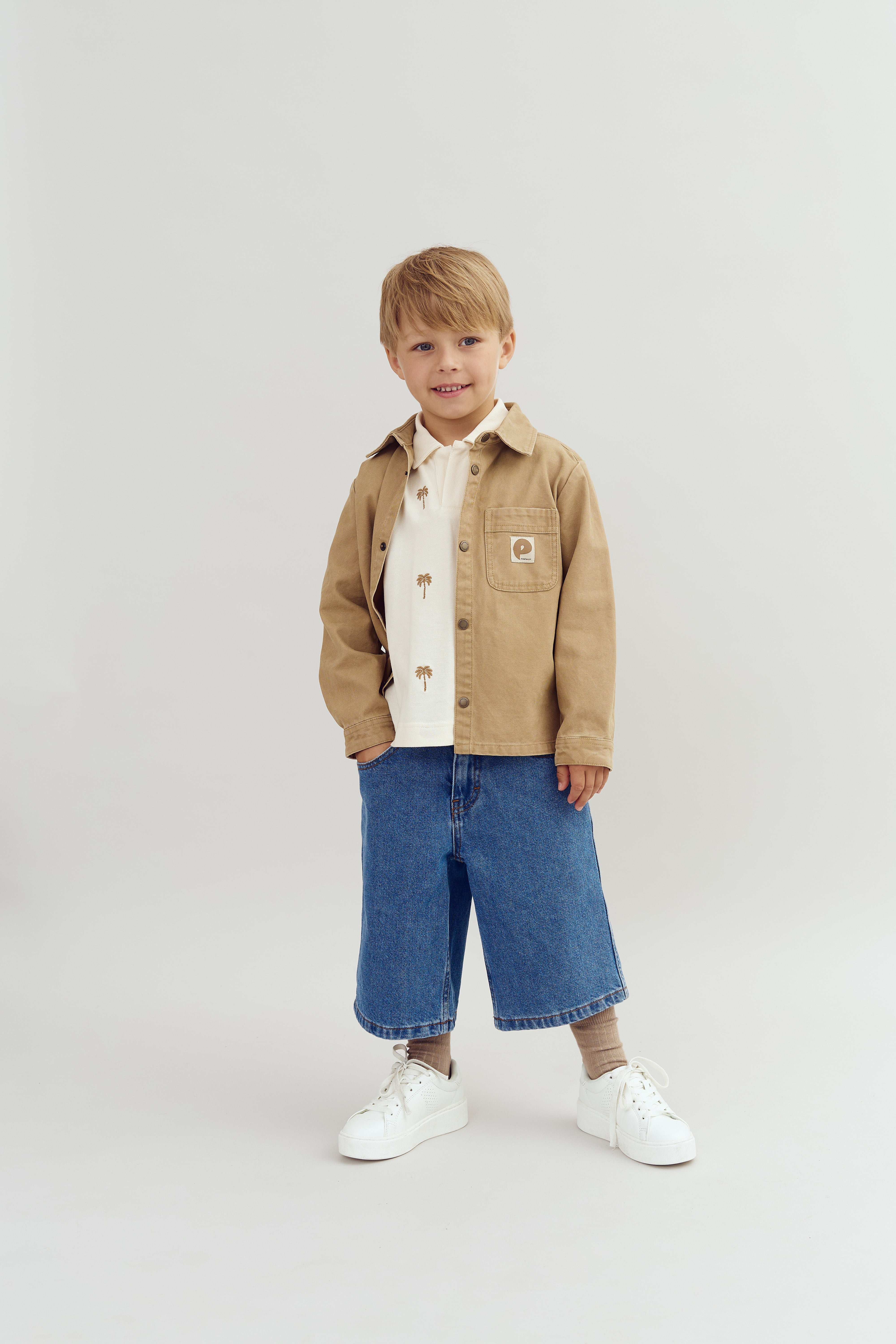 Dreng står og smiler iført en beige jakke, hvid skjorte med palmetræer og blå jeansshorts. Baggrunden er enkel og lys, hvilket fremhæver hans tøjstil.