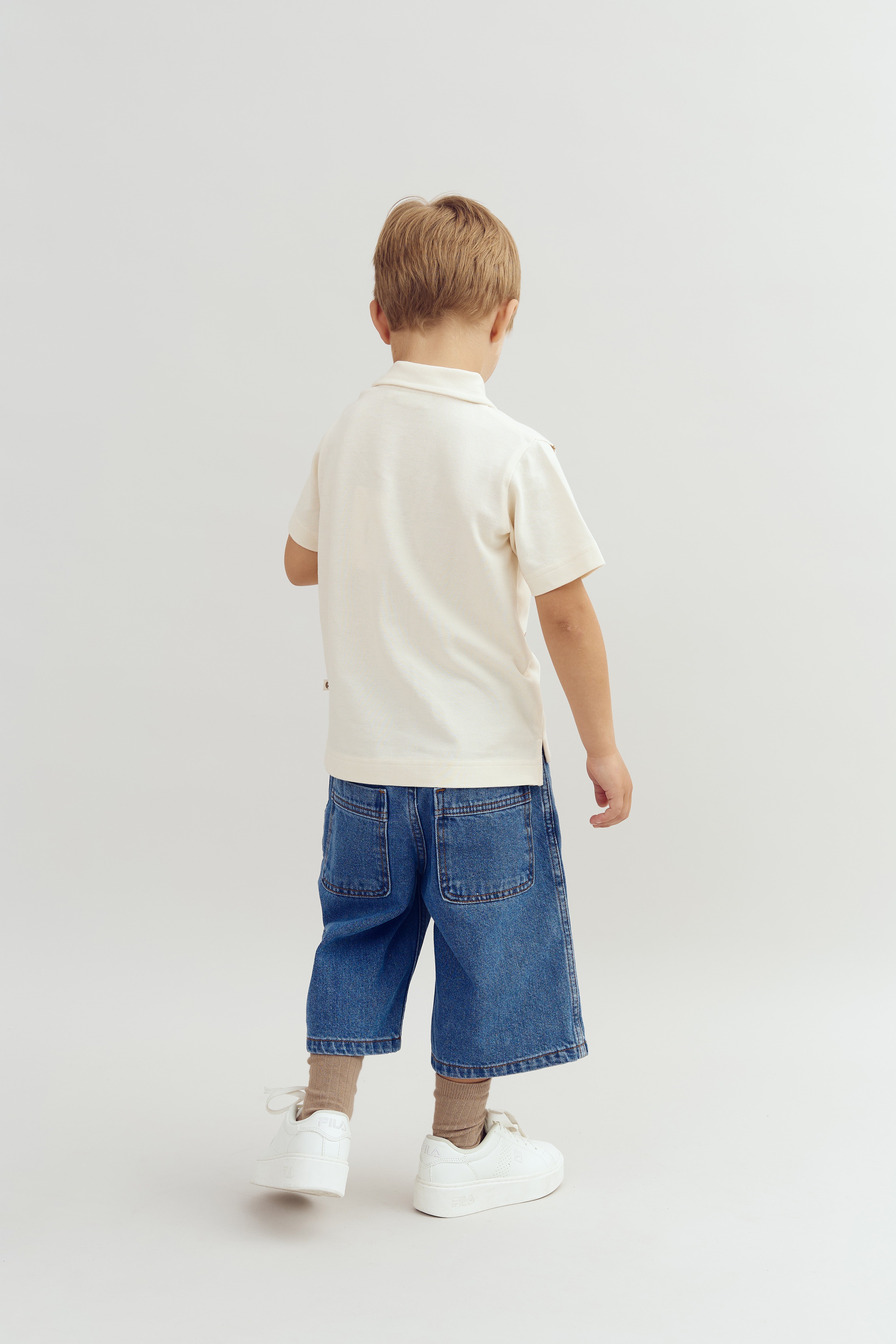 En dreng står med ryggen til, iført en cremefarvet trøje og blå shorts, på en lys baggrund. Han har hvide sneakers og brune strømper.