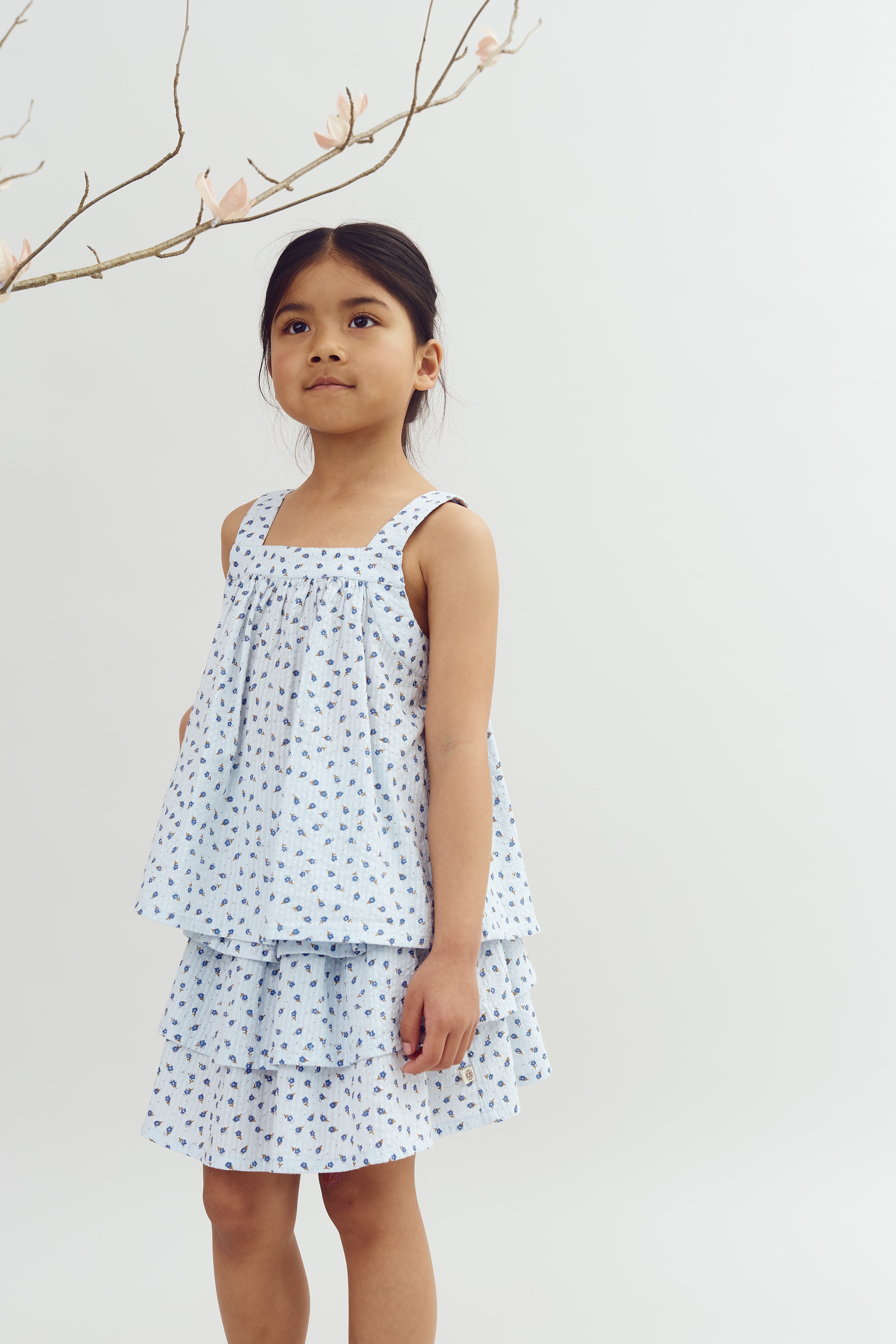 A young girl stands looking upward, wearing a light blue floral dress, with branches and light pink blossoms above, set against a plain white background.