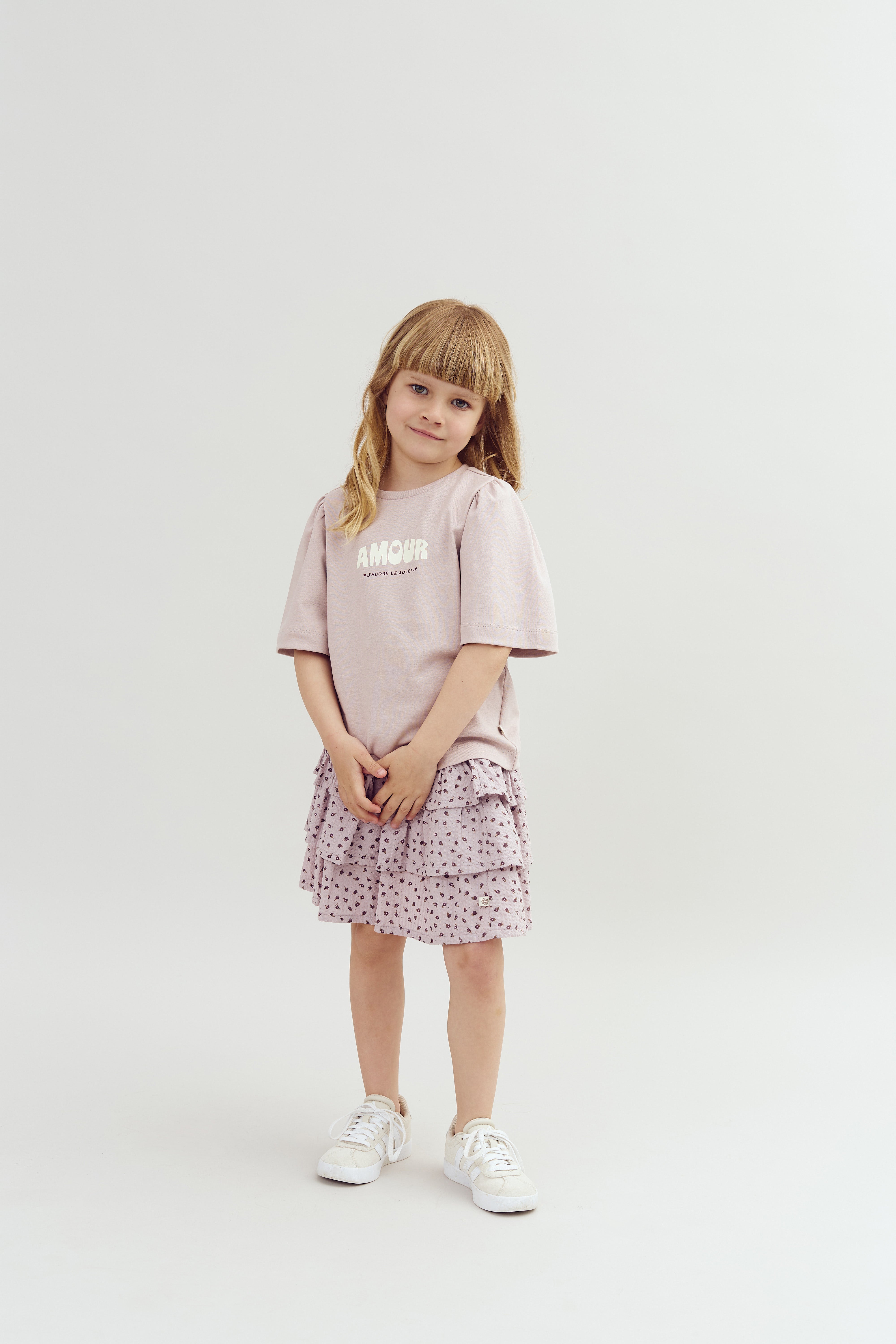 A young child stands in a studio setting wearing a pink shirt with "AMOUR" and "L'AMOUR LE PLUS" printed on it and a pink, patterned skirt, paired with white sneakers.