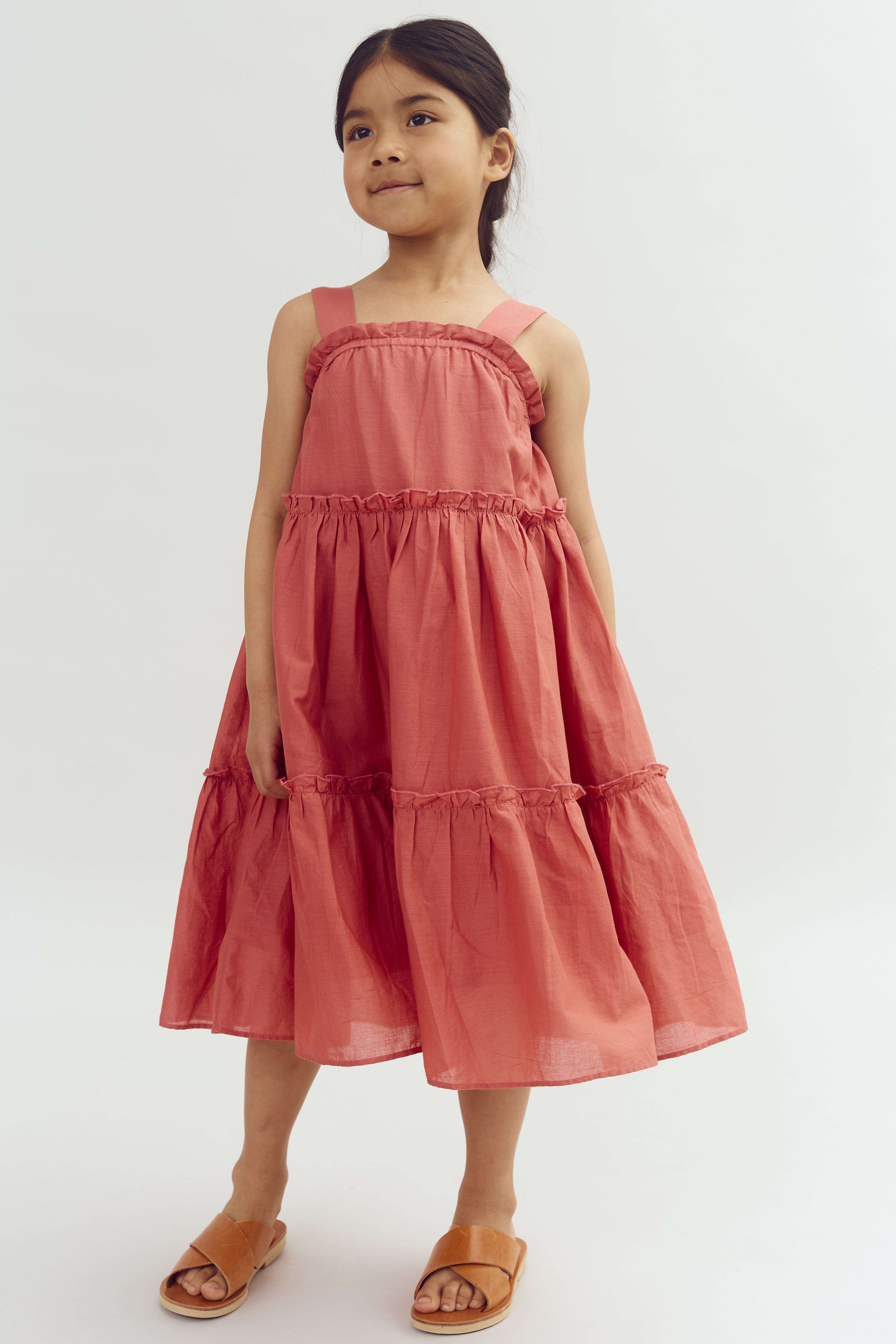 A young girl stands posing, wearing a sleeveless, tiered coral dress and brown sandals in a neutral studio setting.