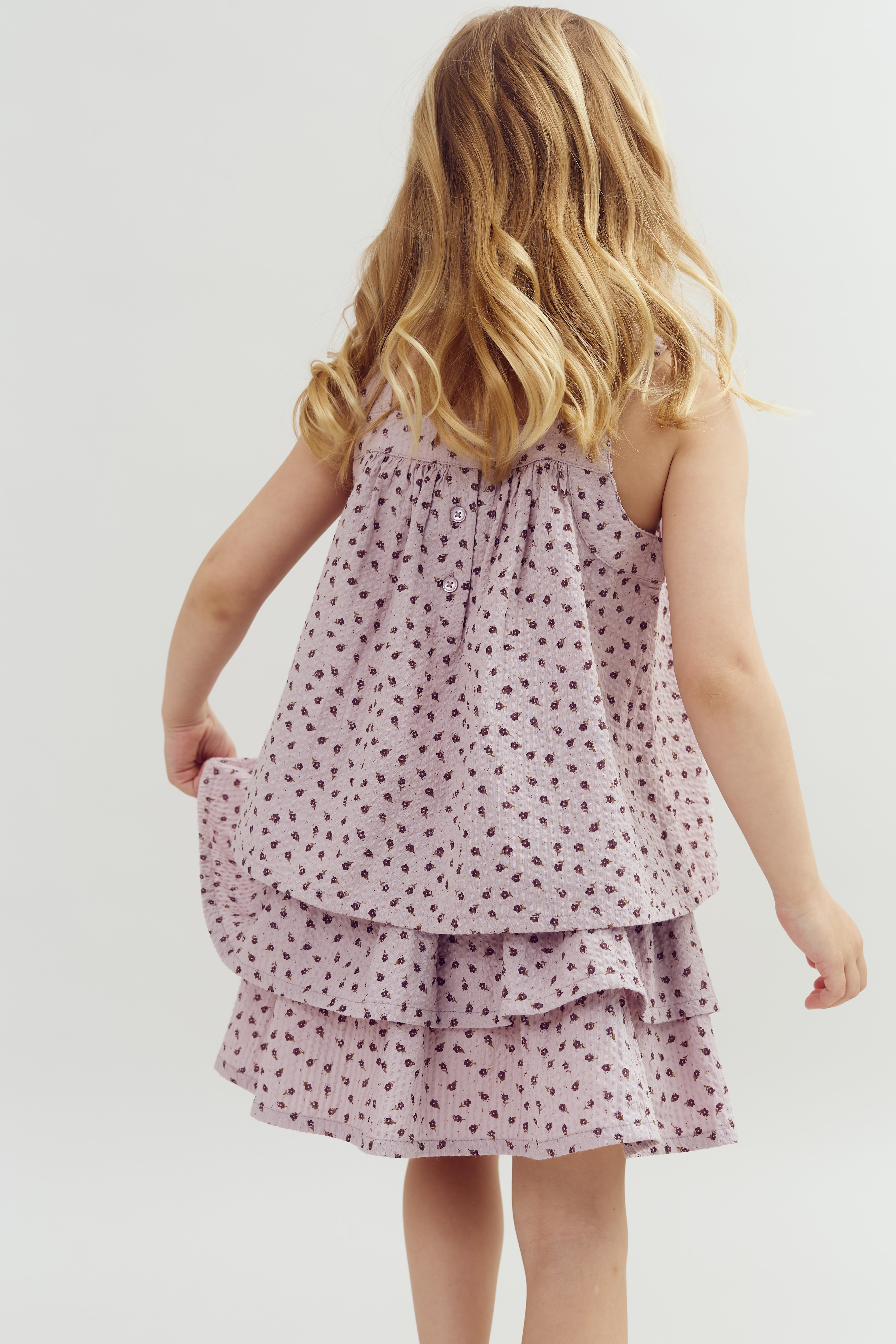 A child with long, wavy hair wears a floral-patterned dress, holding its skirt, standing in a plain, light-colored background.