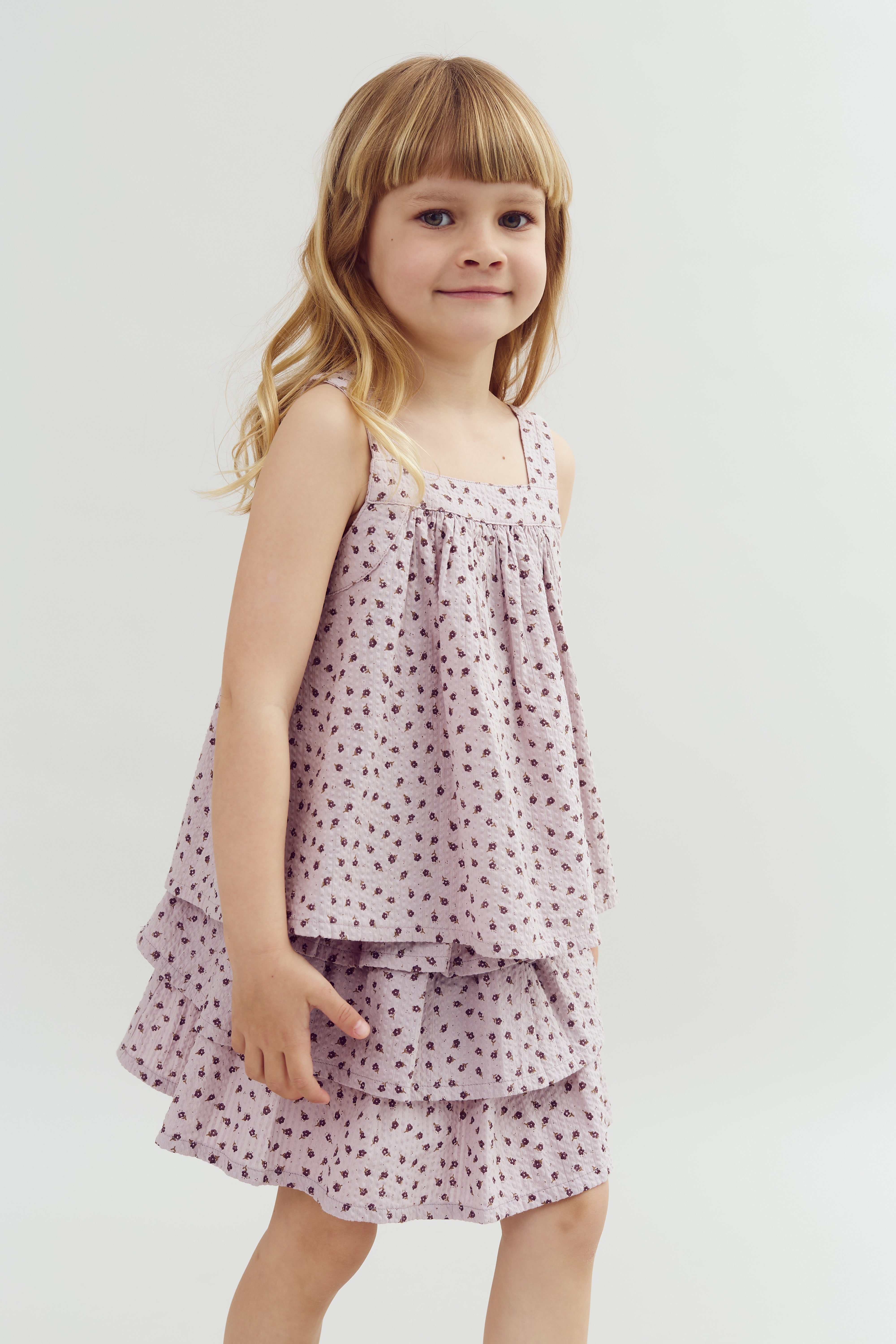 A young child with long hair wears a sleeveless, ruffled dress with a small floral pattern, standing against a plain, light background.