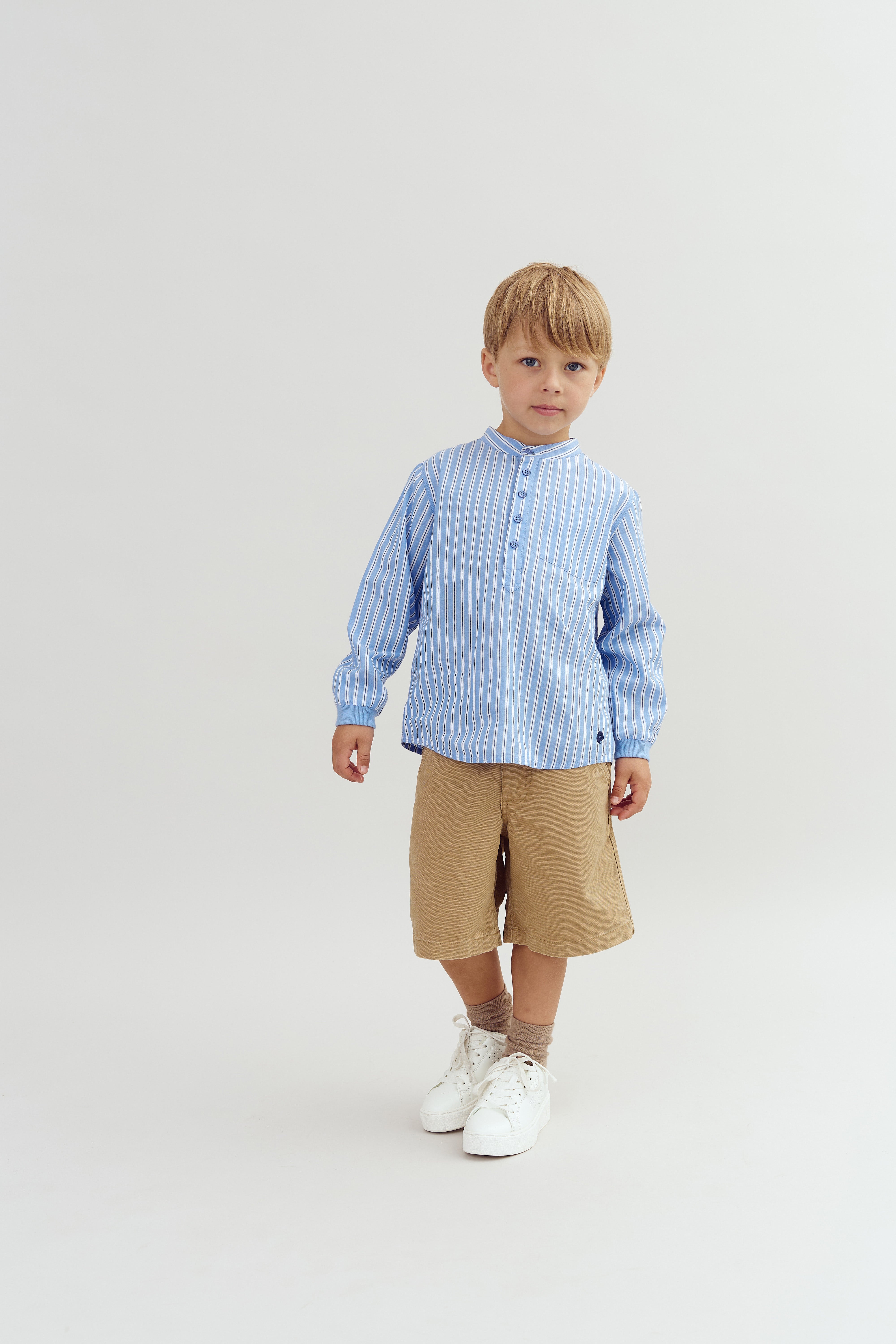 En dreng iført en blåstribet skjorte og beige shorts står oprejst i et neutralt, lyst studie. Han har hvide sneakers på og ser roligt mod kameraet.
