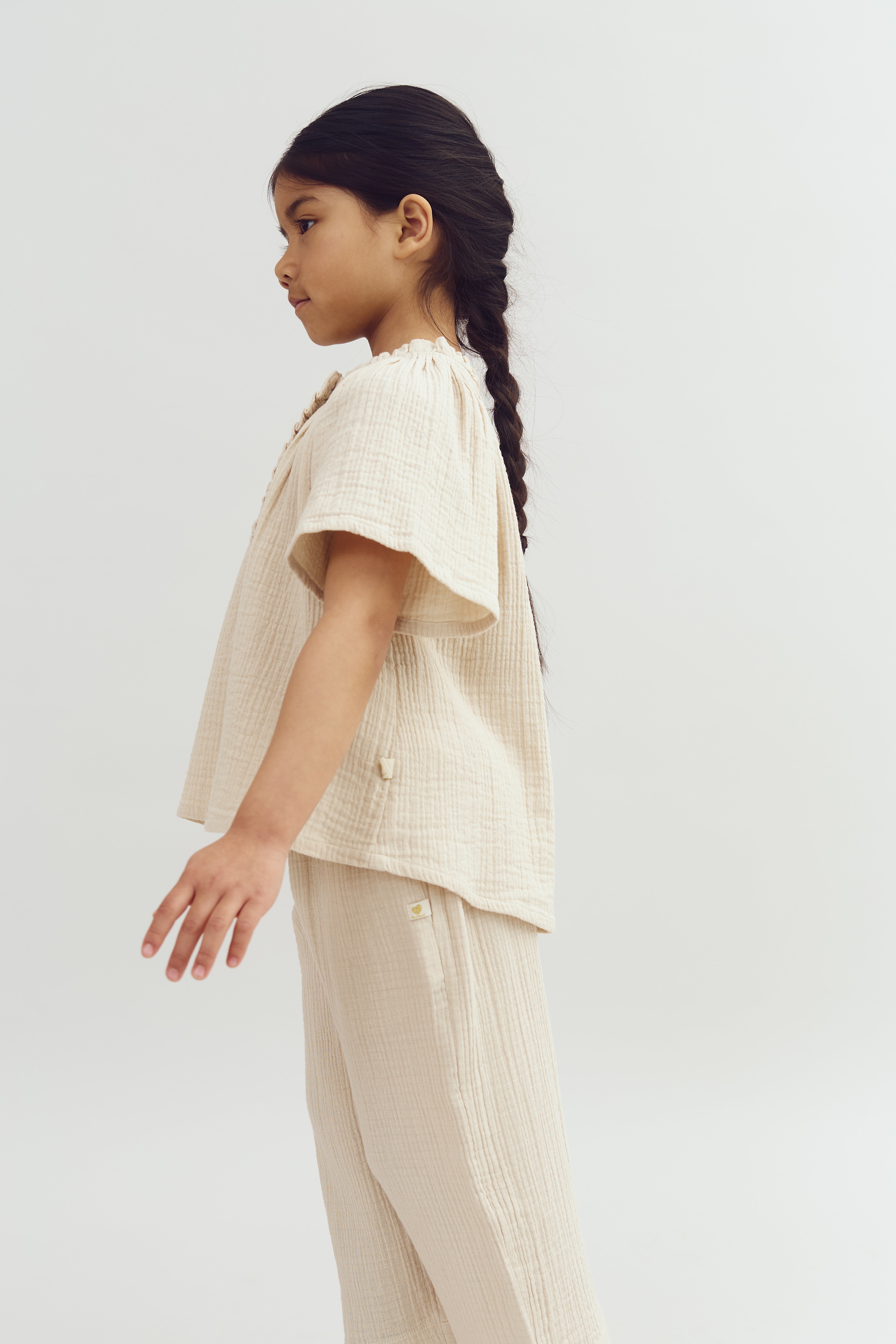 A child stands sideways, wearing a light, textured, cream outfit. They have long, dark hair in a braid, against a plain white background.