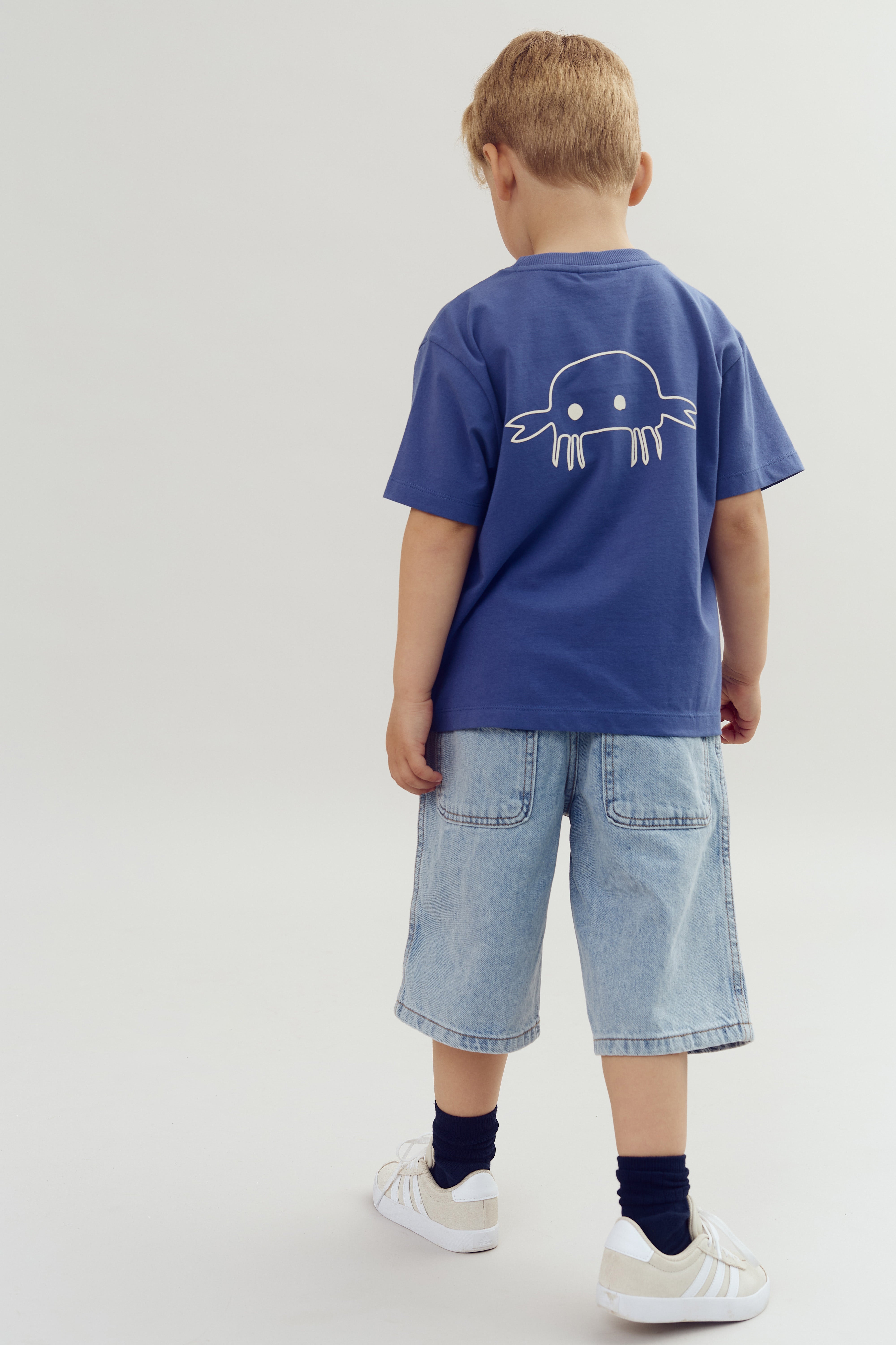 Et barn står med ryggen til iført en blå T-shirt med en hvid krabbe-tegning og denimshorts. Barnet er i et enkelt, lyst rum med hvide sneakers på.
