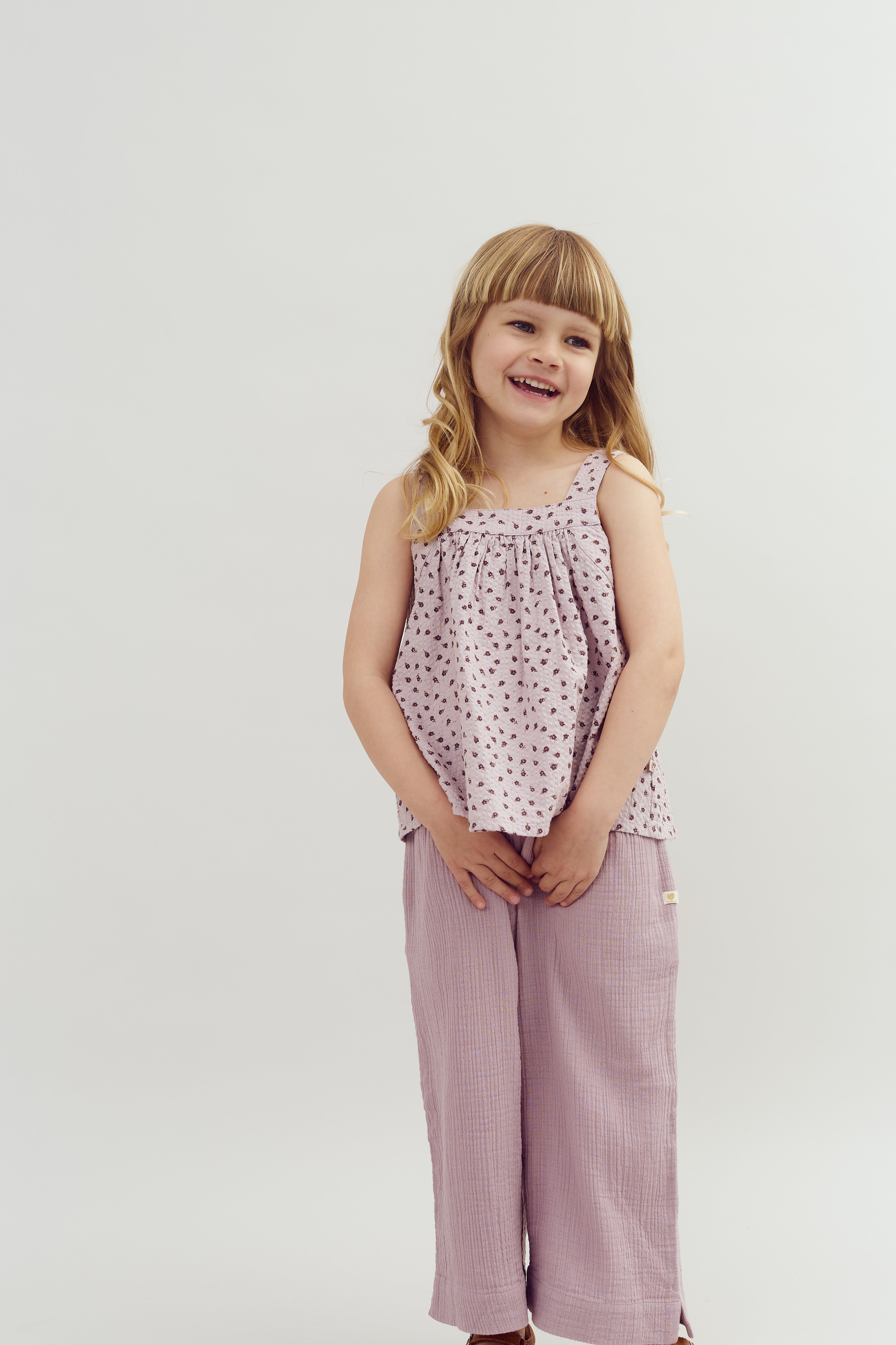 A young girl stands smiling, wearing a sleeveless patterned top and loose lavender pants, hands clasped in front. The background is a plain, light-colored backdrop.