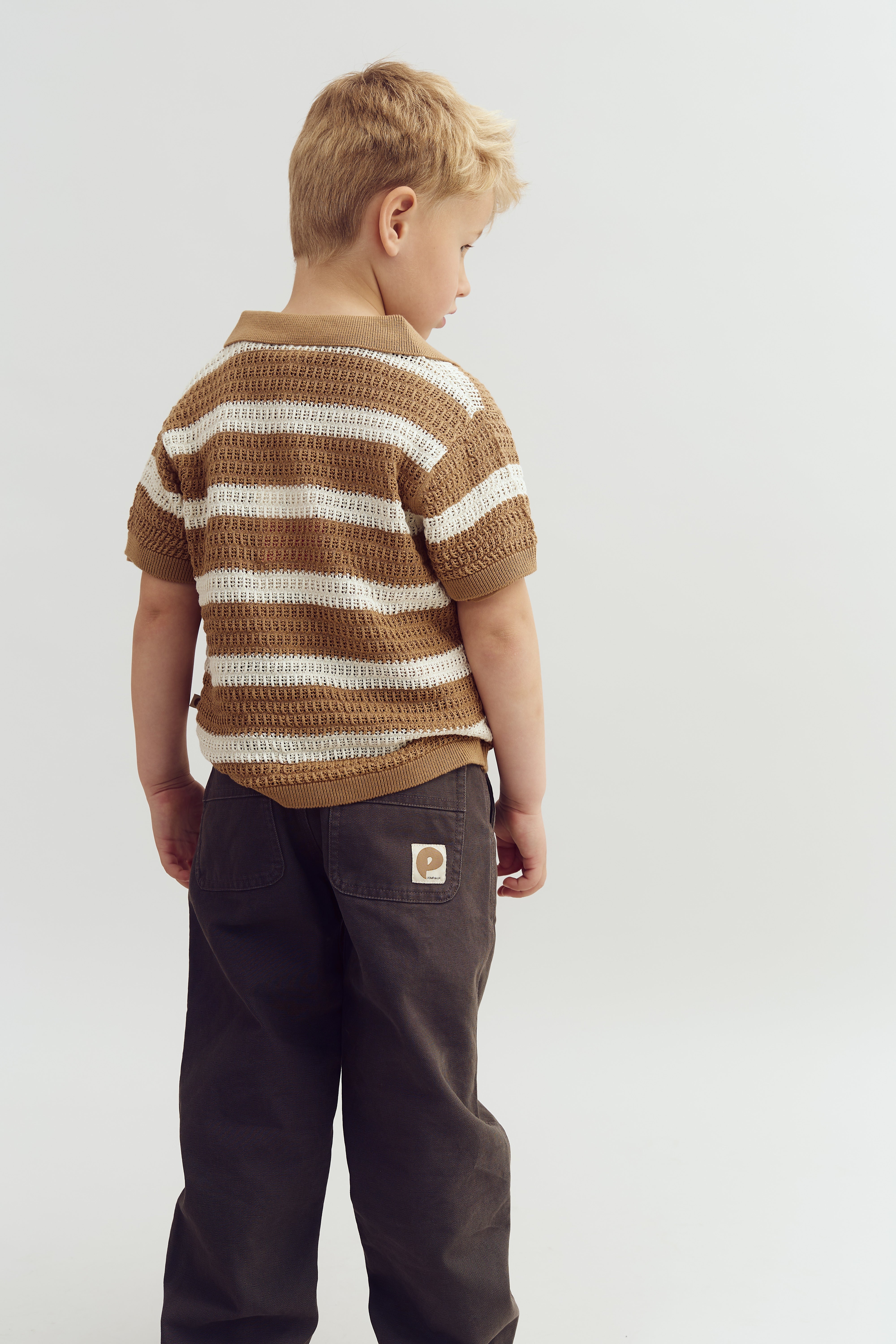 Dreng set fra ryggen, iført en stribet trøje og mørke bukser med et logo. Står mod en neutral, lys baggrund.