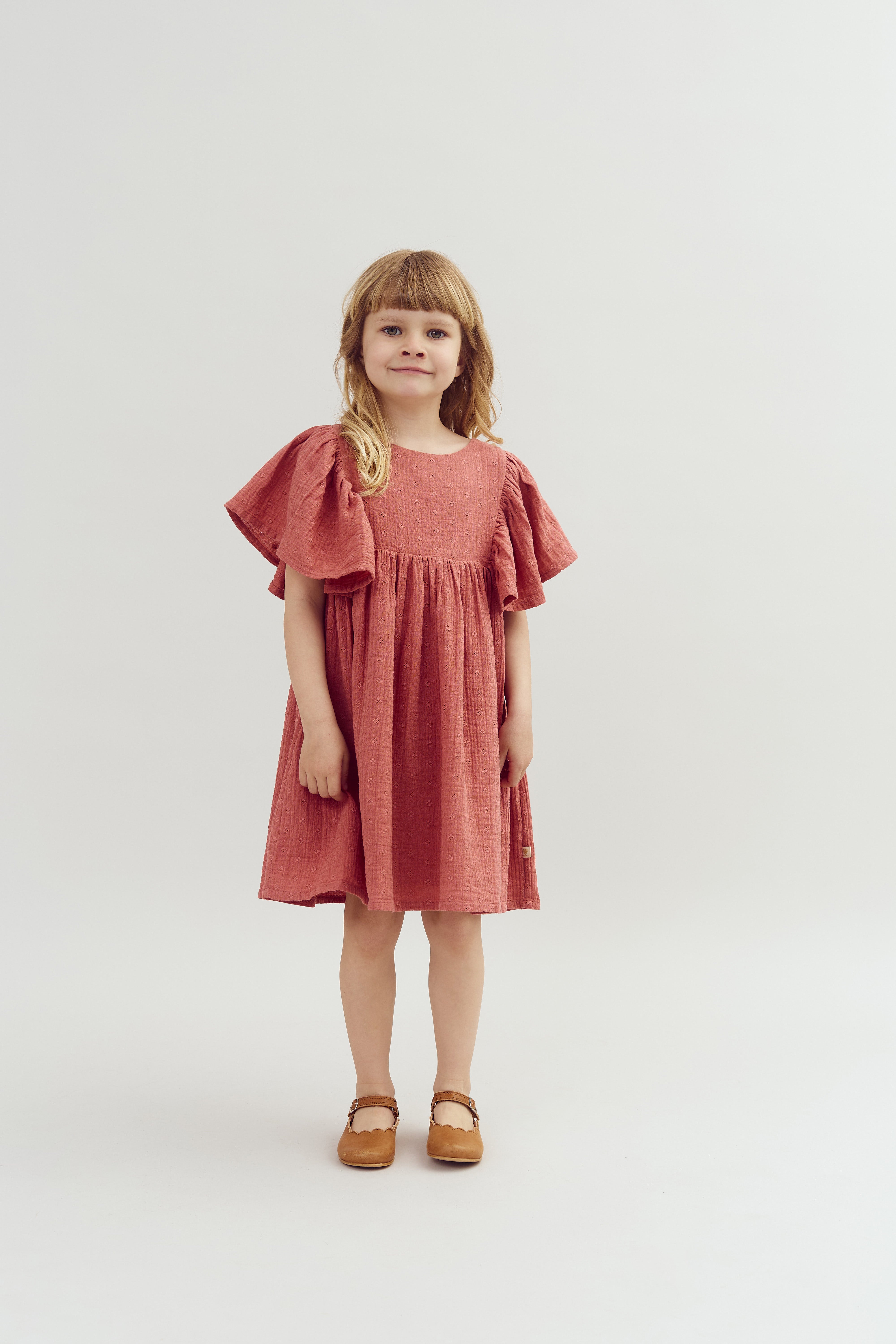 A young girl stands smiling, wearing a flowing, rust-colored dress with ruffled sleeves and brown shoes, set against a plain light background.
