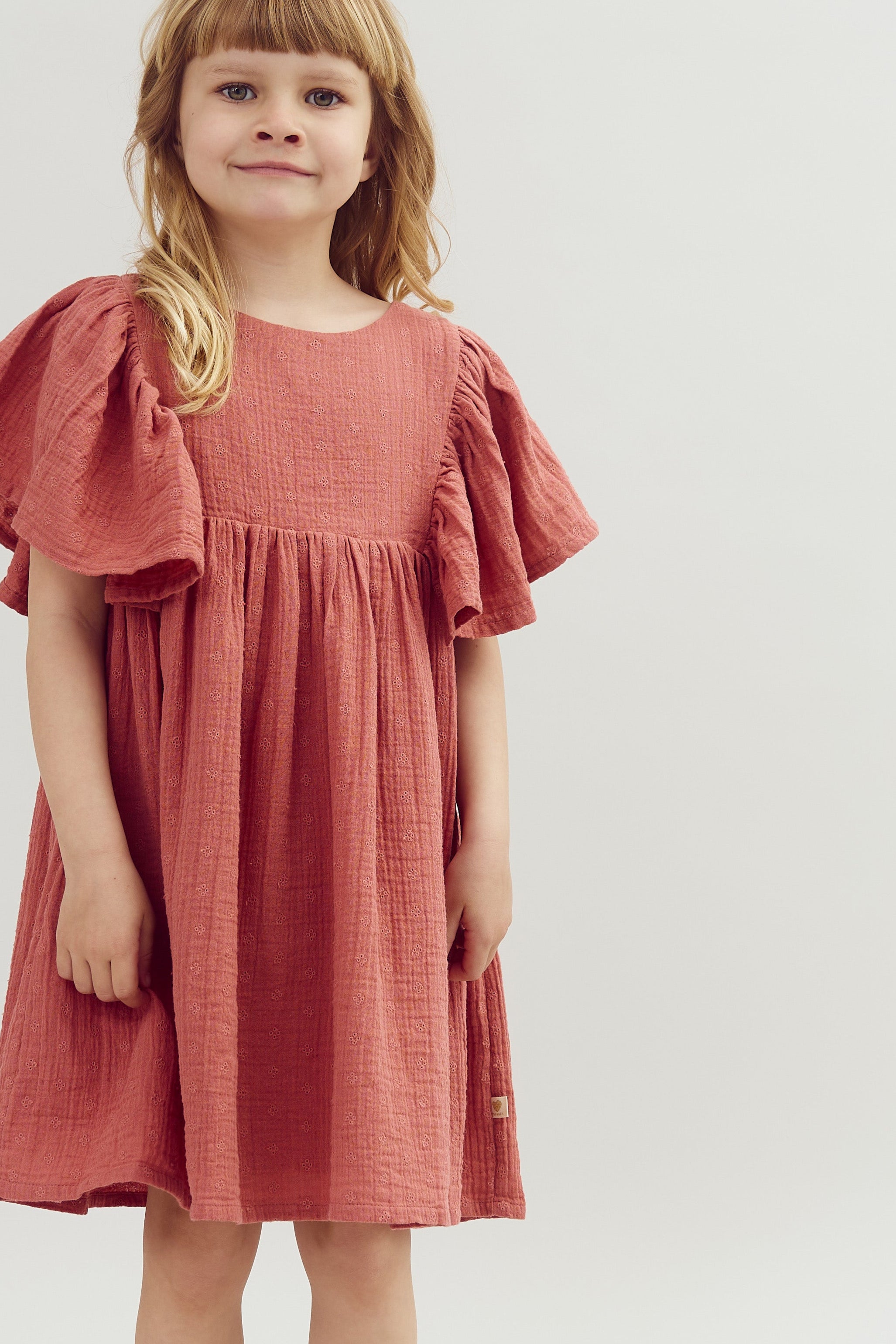 A child stands smiling, wearing a red dress with flared sleeves, in front of a plain light gray background.
