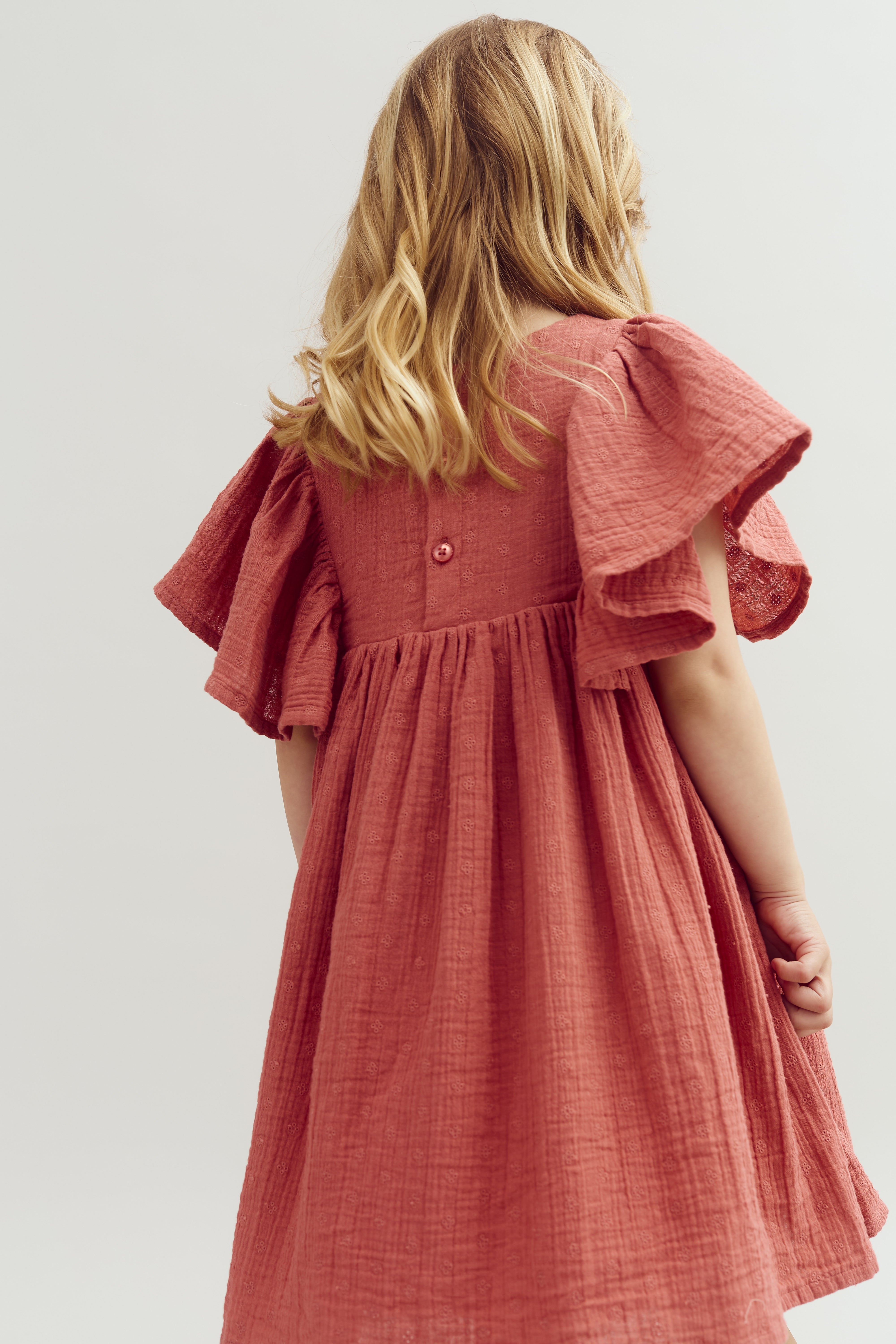 A young child with blonde hair stands slightly turned, wearing a textured, pink dress with flowing sleeves, in a neutral setting.
