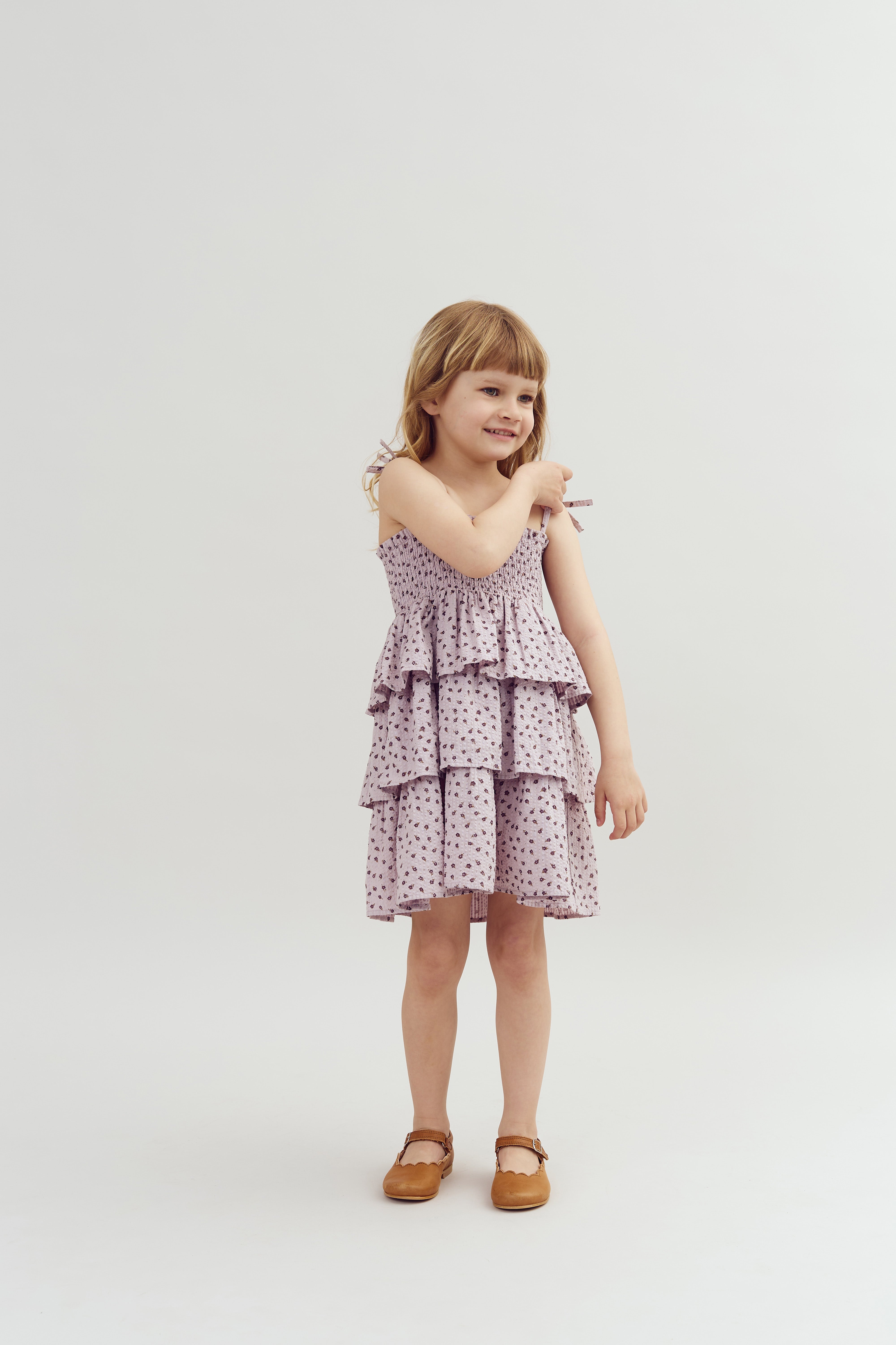A young girl stands smiling, wearing a tiered, patterned dress and brown shoes, against a plain white background.