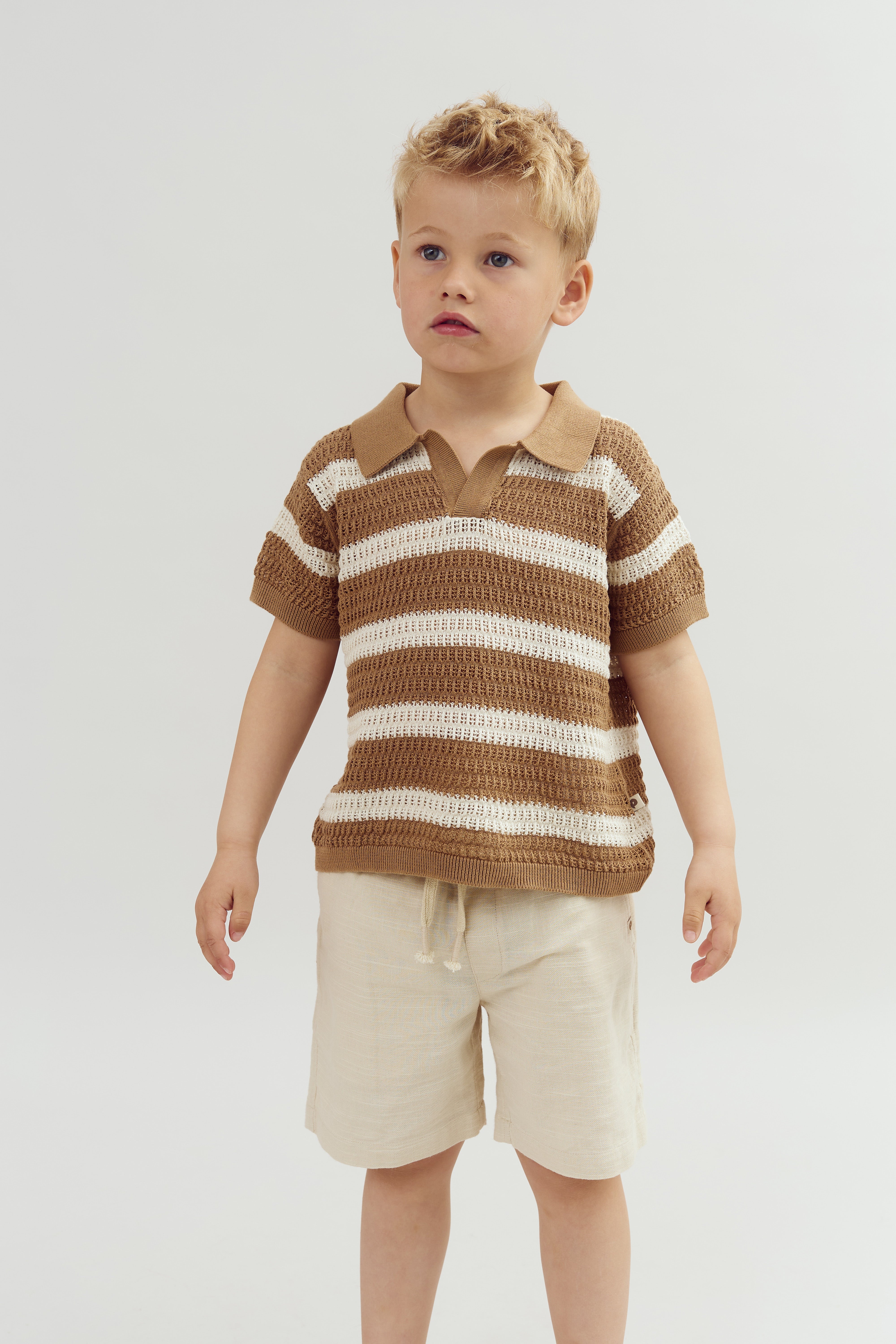 Child stands wearing a brown and white striped shirt and beige shorts in a neutral indoor setting, looking attentively to the side.