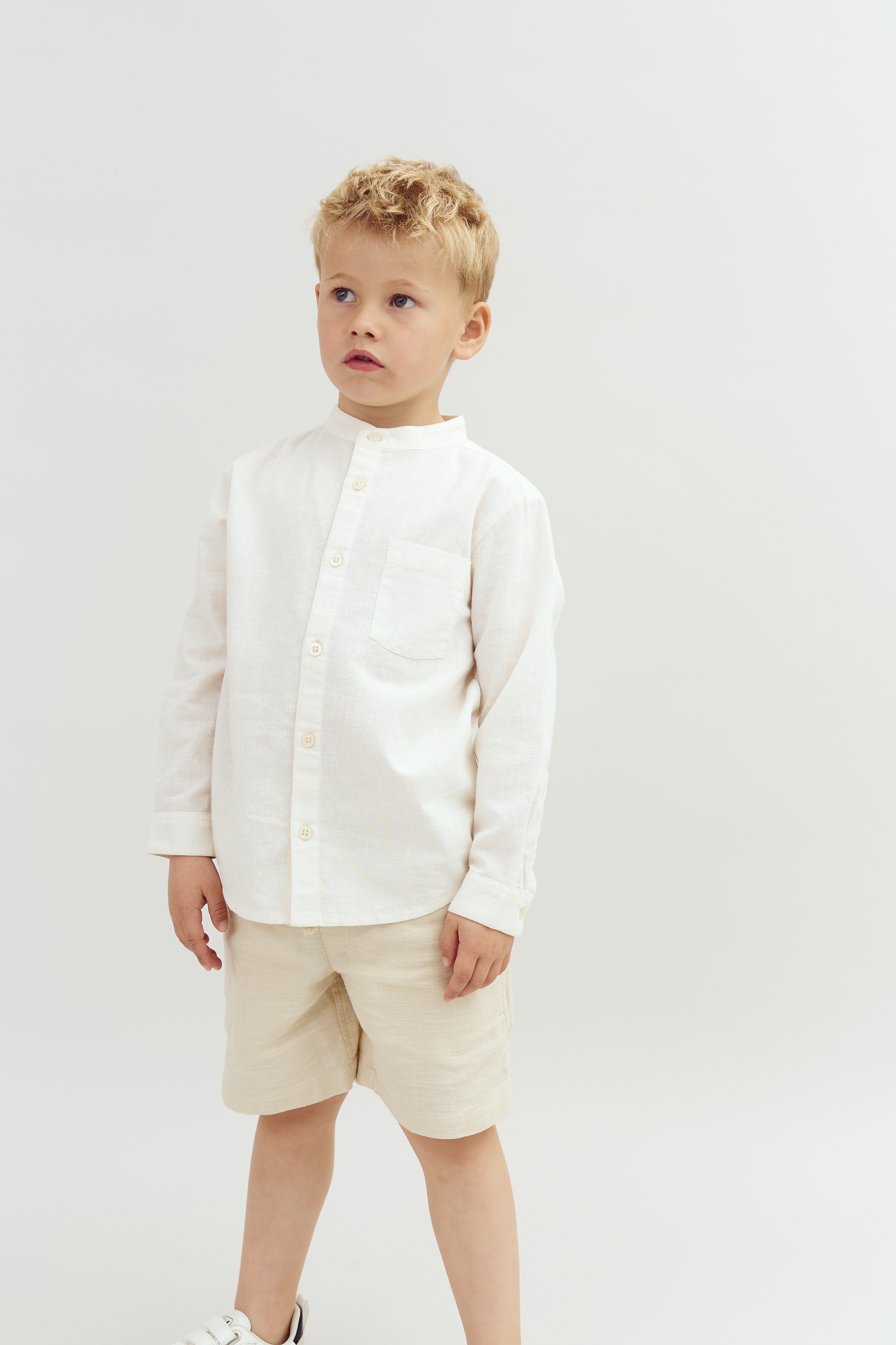 En dreng står stille i beige shorts og hvid skjorte mod en ensfarvet baggrund. Han ser til venstre med et fokuseret udtryk.