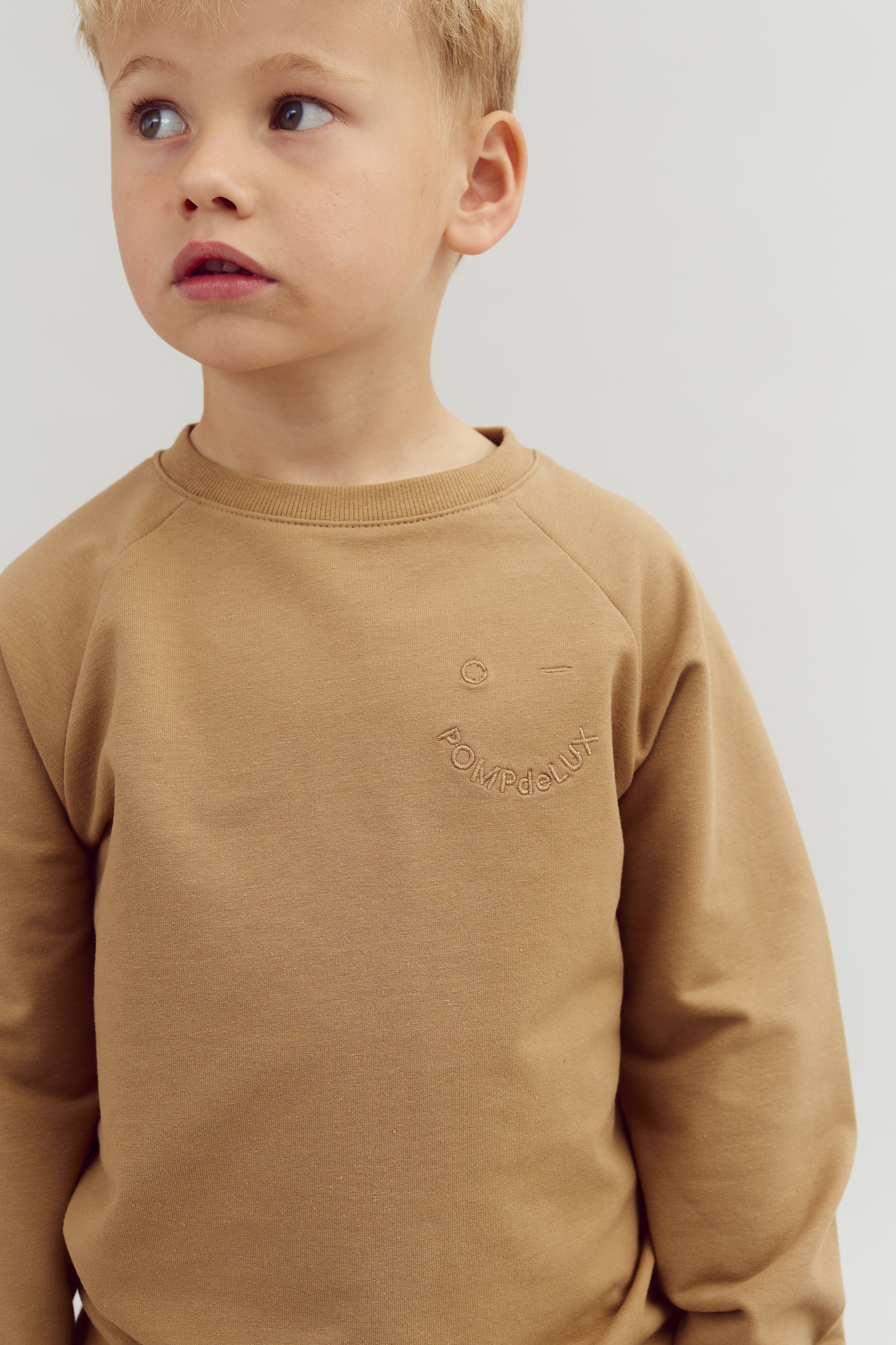En dreng i en brun sweatshirt med teksten "POMPdeLUX" kigger til siden i et neutralt, lyst rum.
