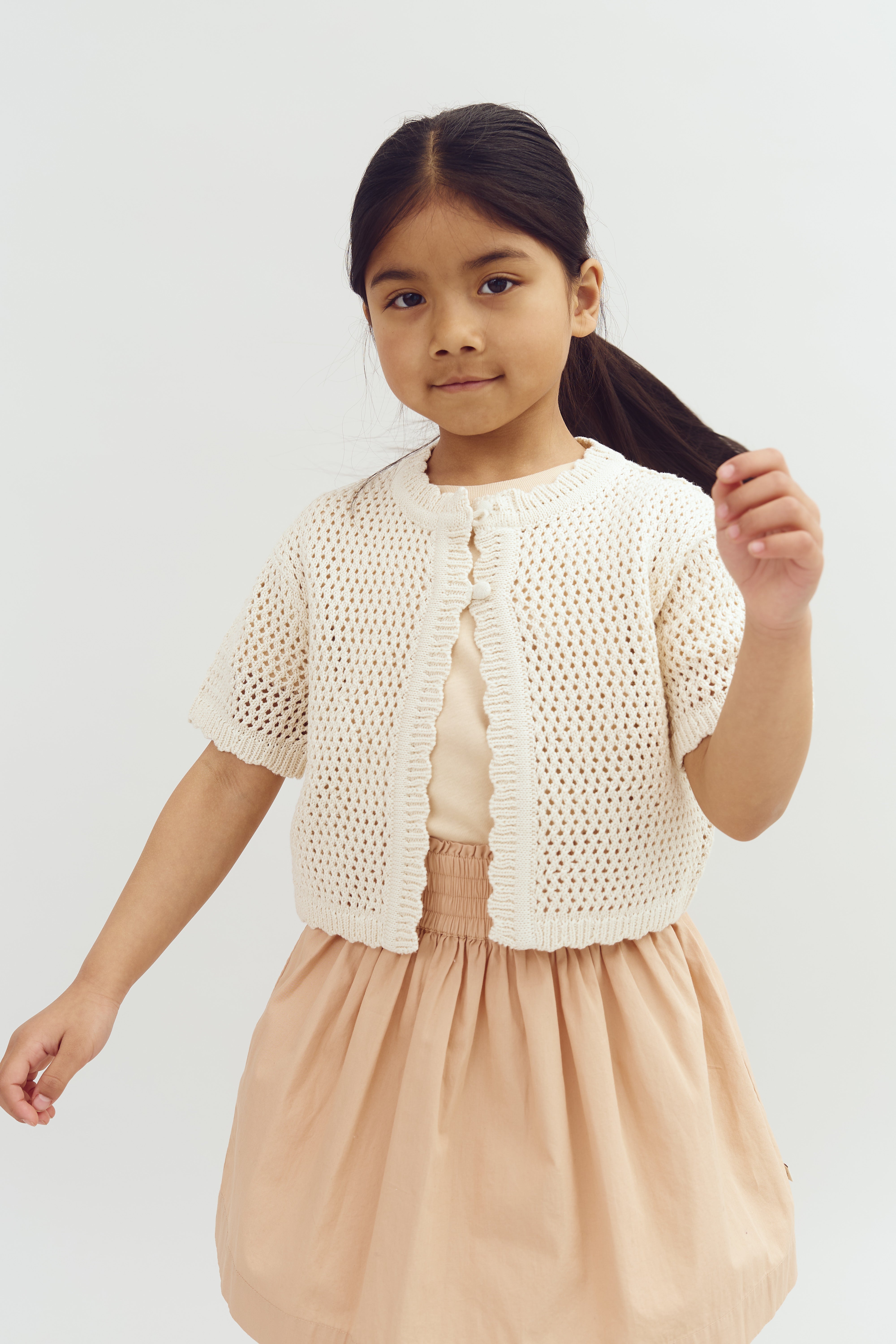 A young girl wearing a knitted cream cardigan over a peach blouse and skirt stands against a plain white background, holding her hair with one hand.