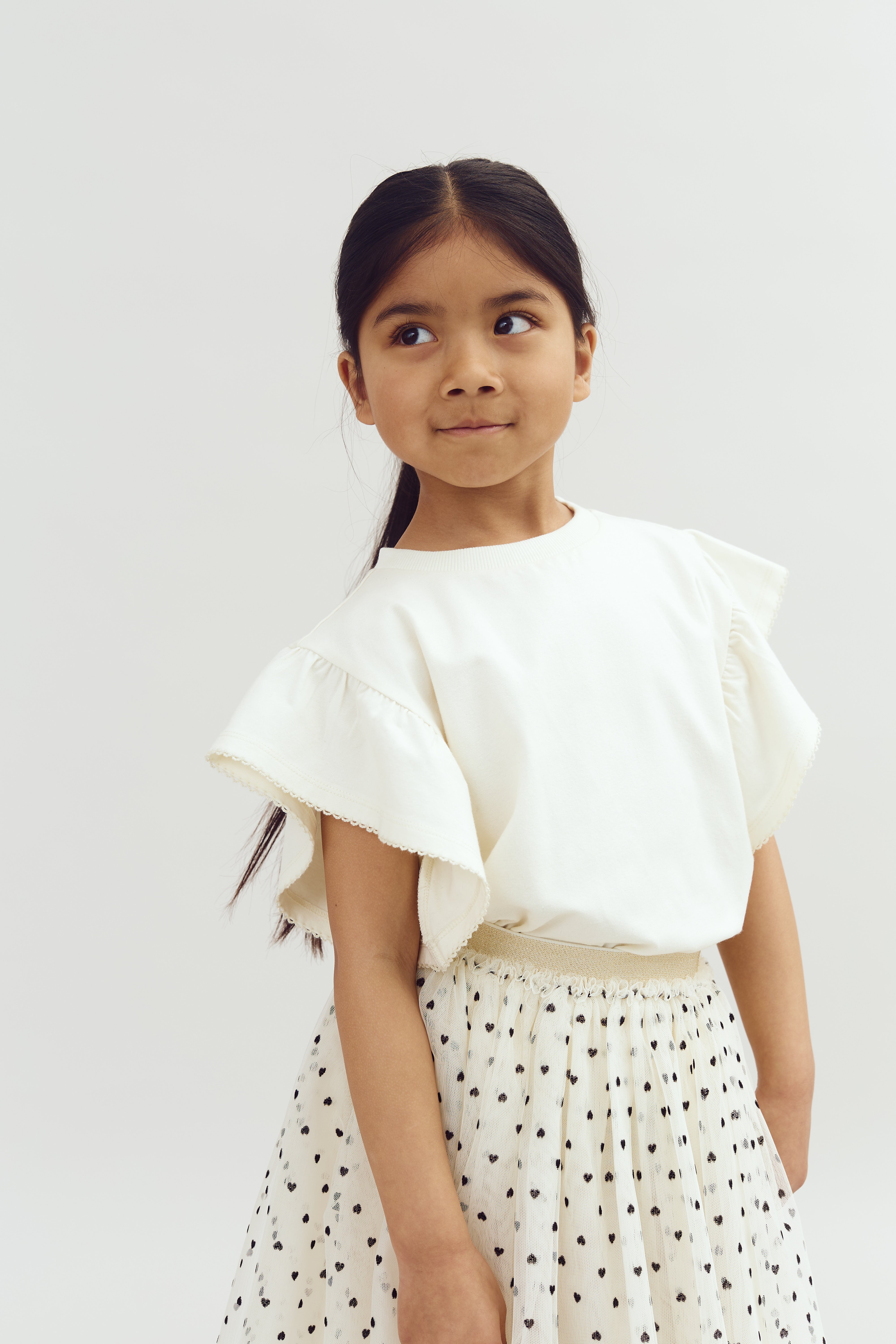 A young girl with long hair, wearing a white ruffled shirt and polka-dot skirt, stands gazing sideways against a plain white background.