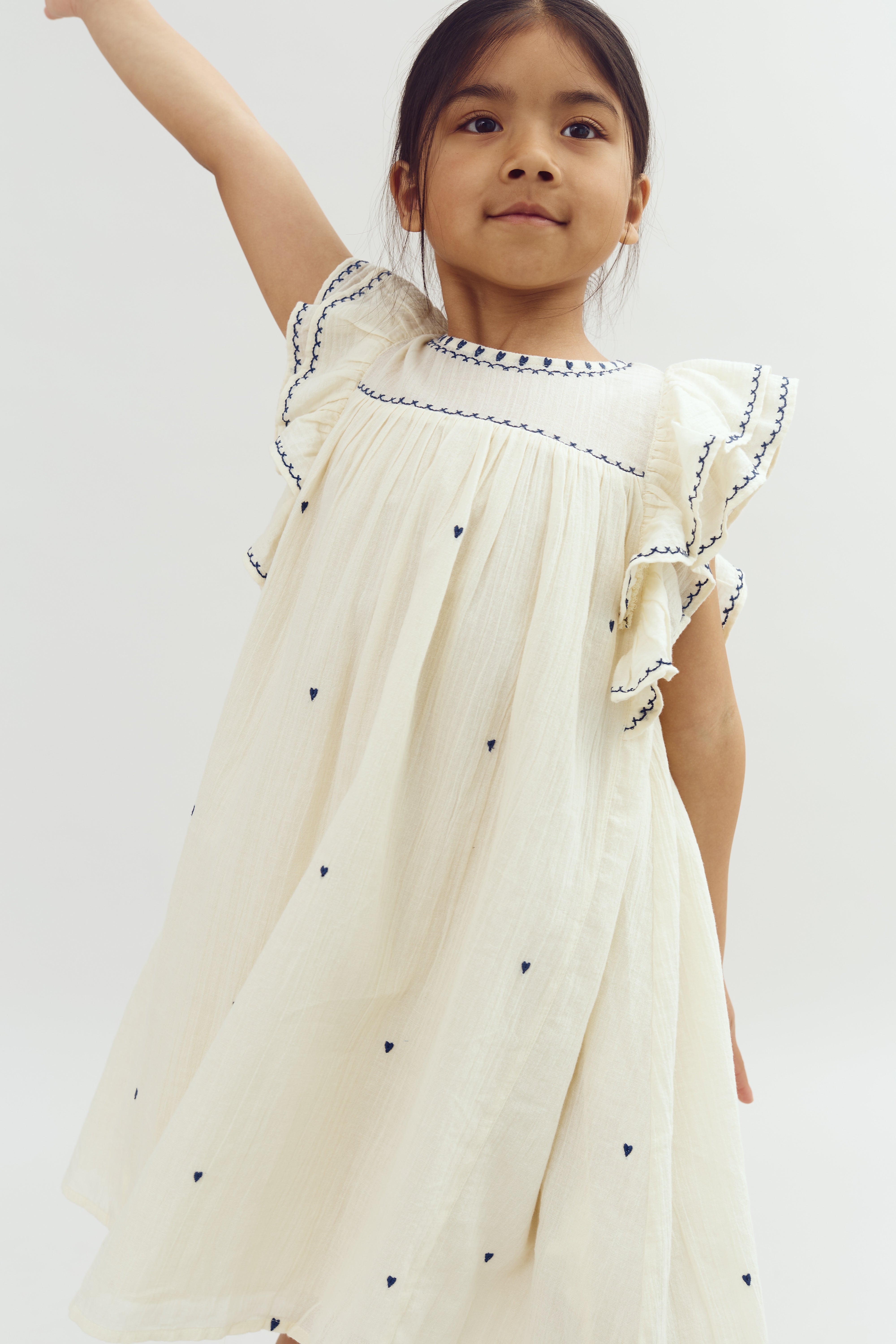 A child wearing a cream dress with navy embroidery and small heart patterns raises one arm, smiling in a neutral, well-lit environment.