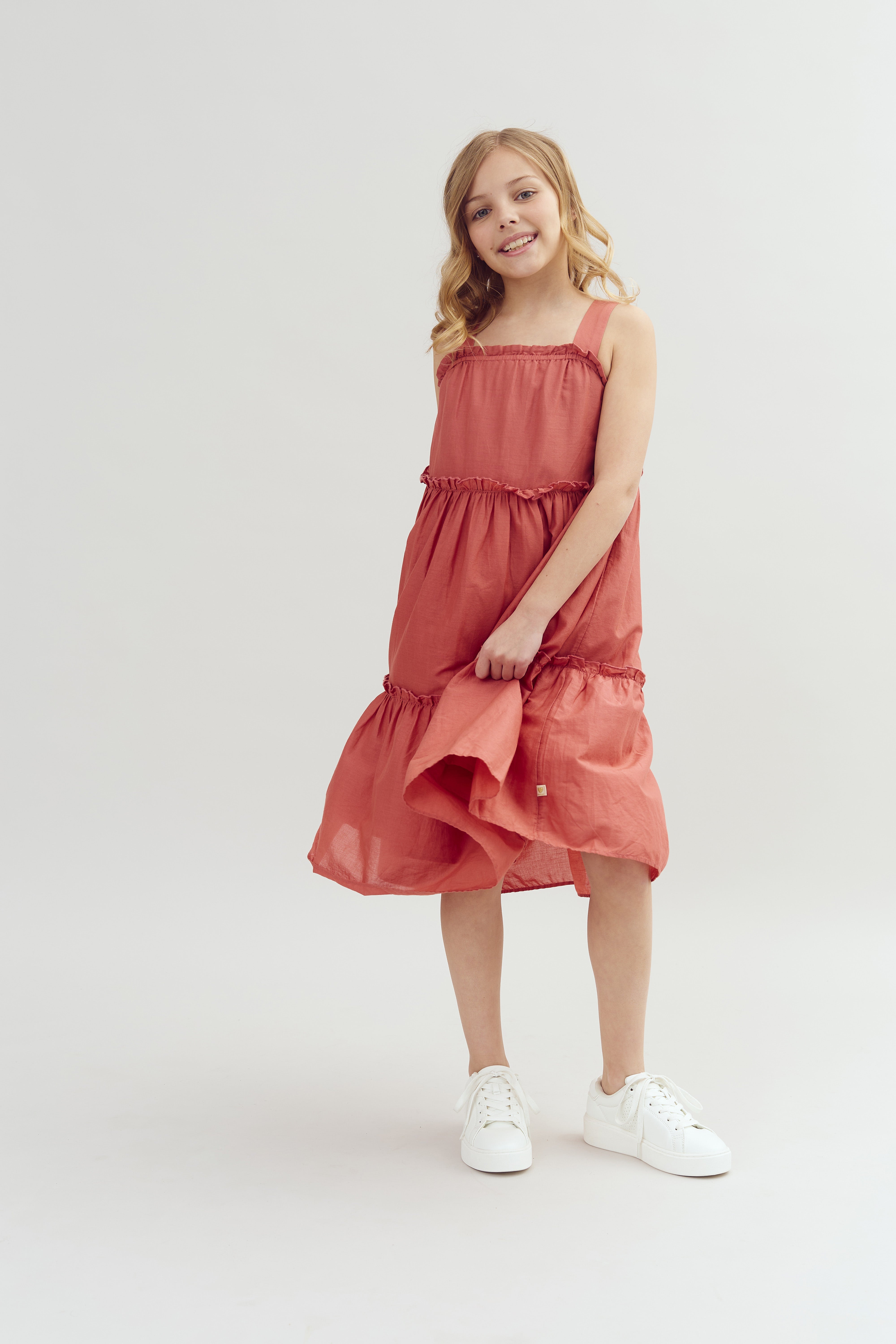 A young girl in a sleeveless, coral dress holds one side of it, smiling softly in a bright, plain studio setting. She wears white sneakers.