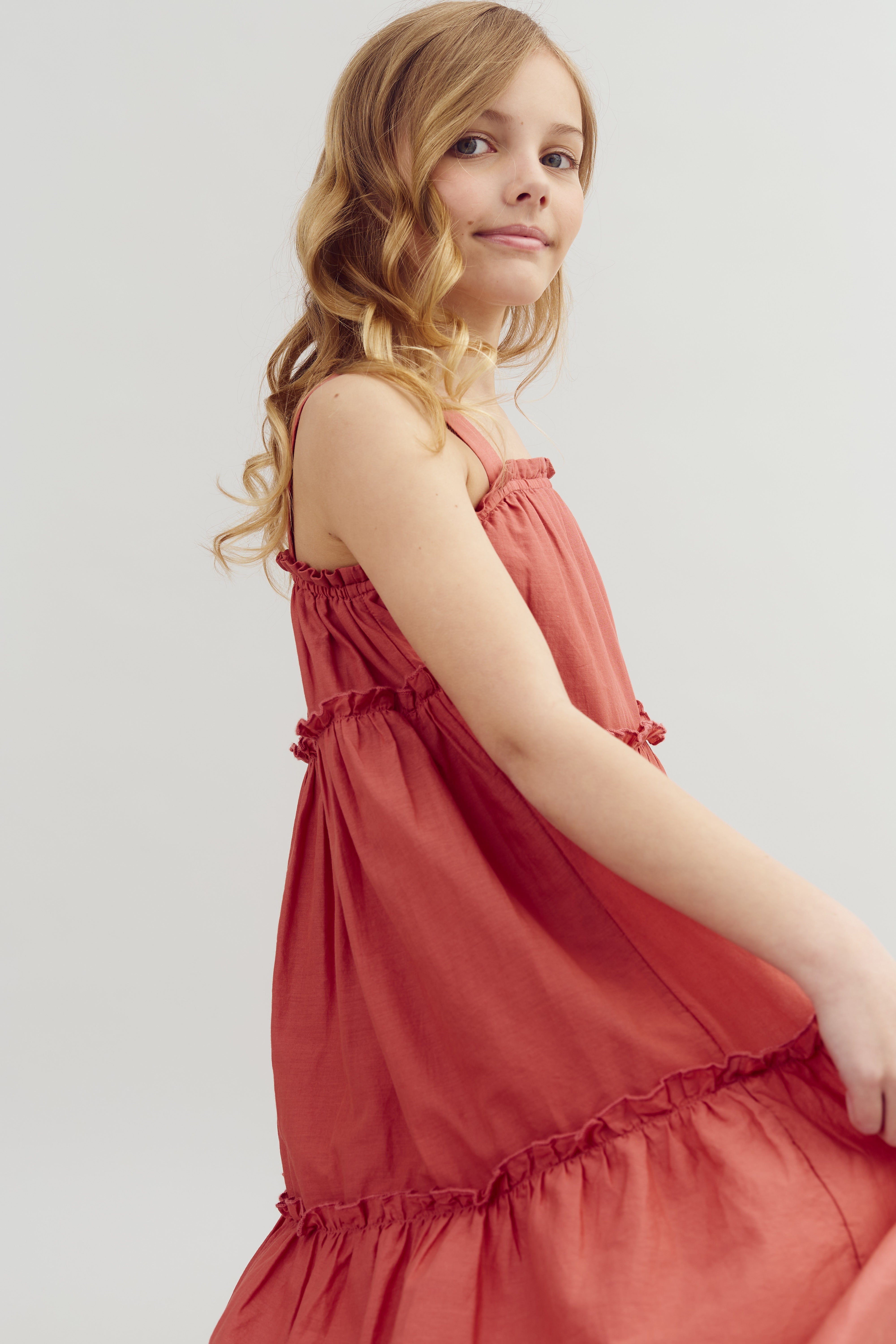 A young girl with curly blonde hair wears a sleeveless red dress, standing against a plain, light-colored background, looking sideways with a soft expression.