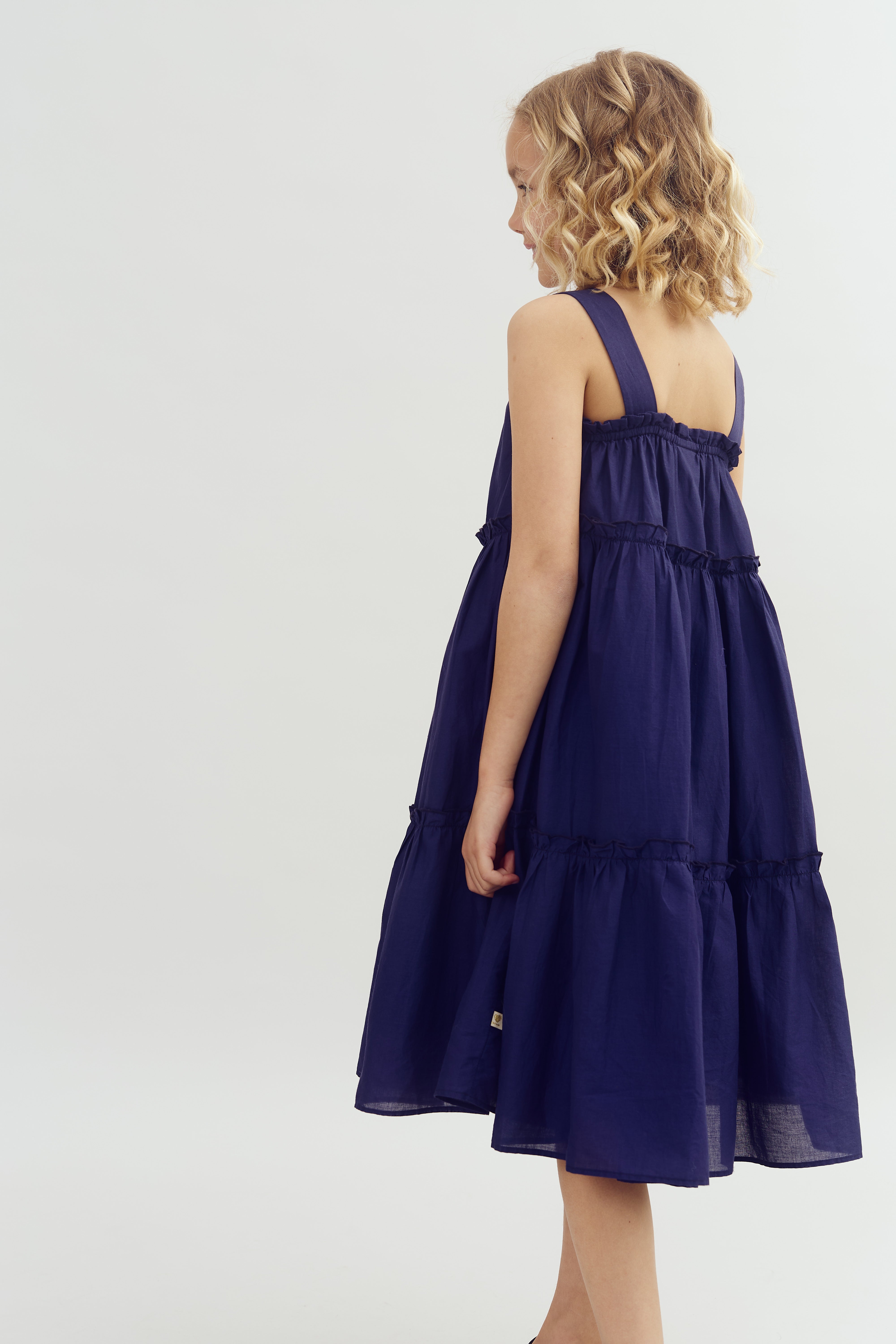 A young girl stands sideways, wearing a navy blue, tiered sundress, with her curly hair draping down. She is in a plain, light-colored studio setting.