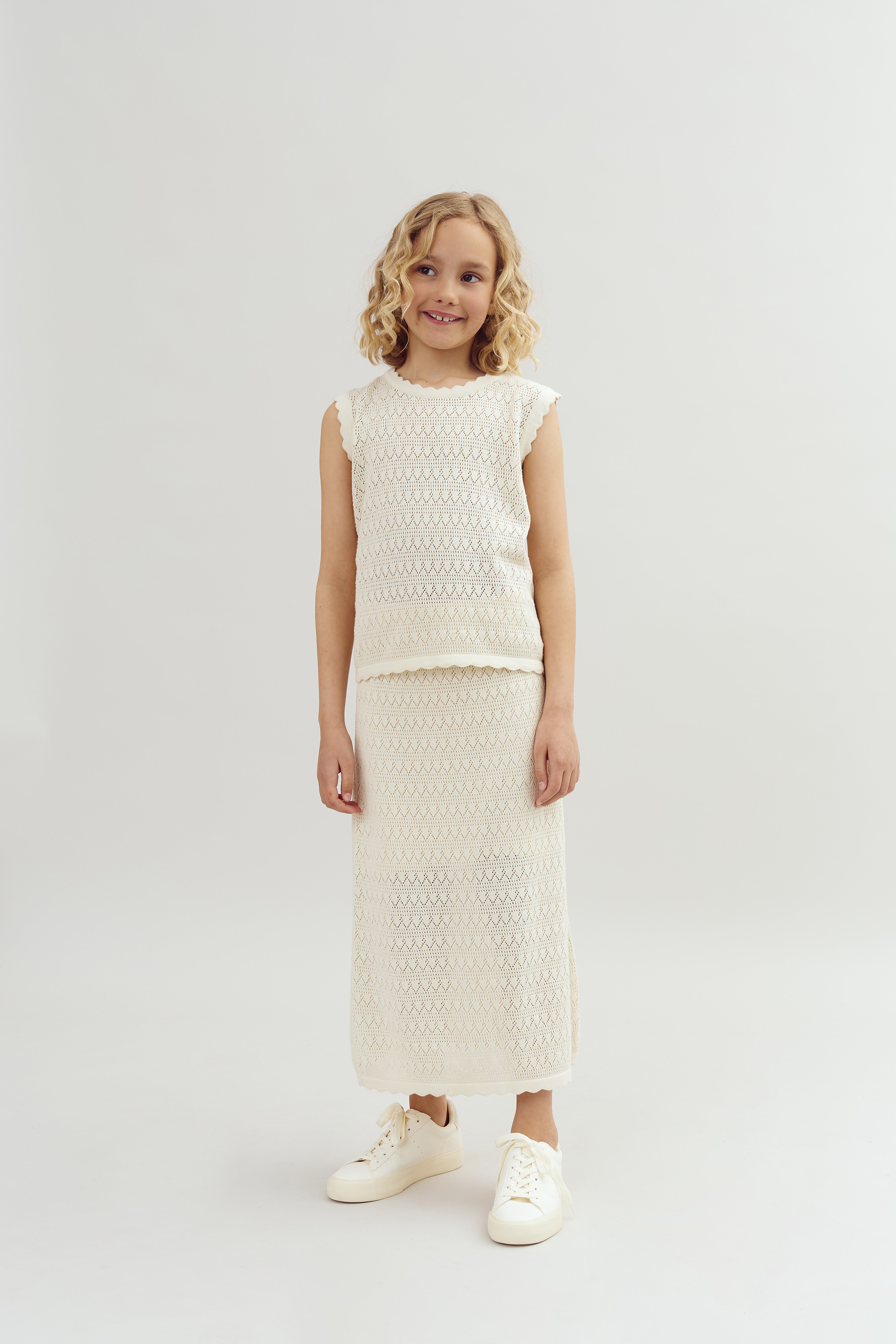 A child wearing a knitted, off-white outfit smiles and stands still, in a plain, studio setting with a light background. She has curly hair and wears matching white sneakers.