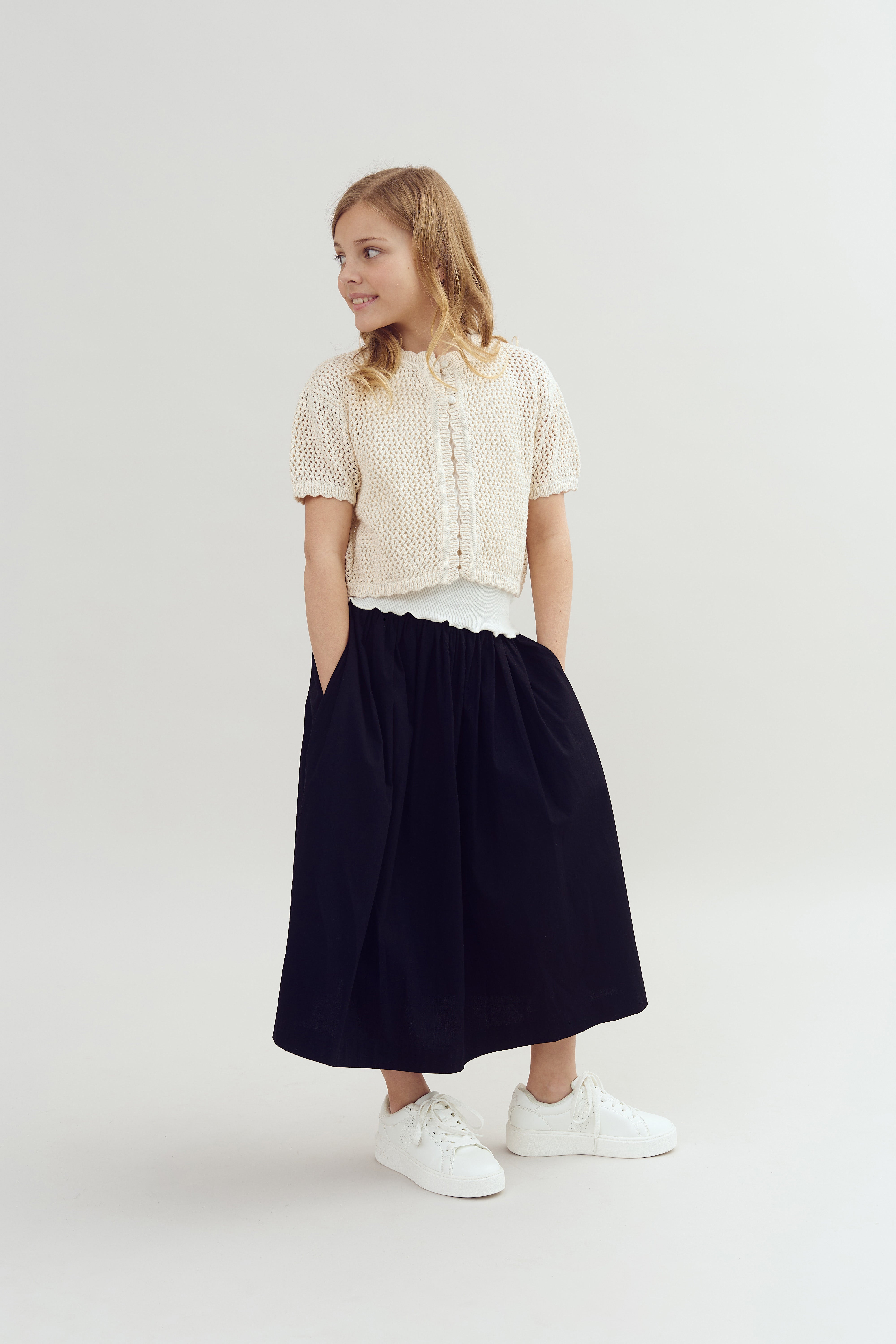 A young girl stands with hands in pockets, wearing a cream knit cardigan, black skirt, and white sneakers, looking to the side against a plain, light gray background.