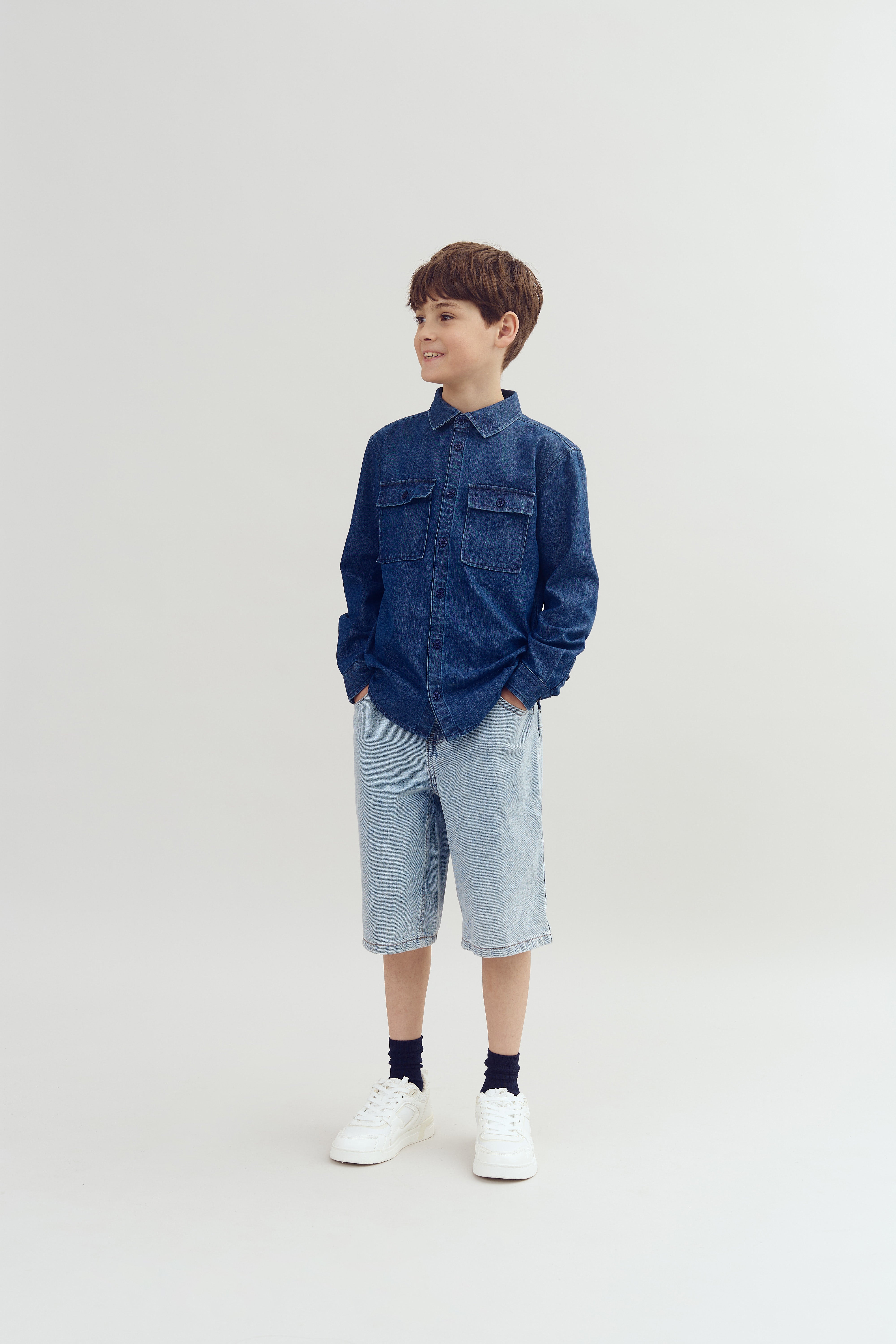 Dreng i denimskjorte og shorts står afslappet med hænderne i lommerne i en lys, neutral baggrund.