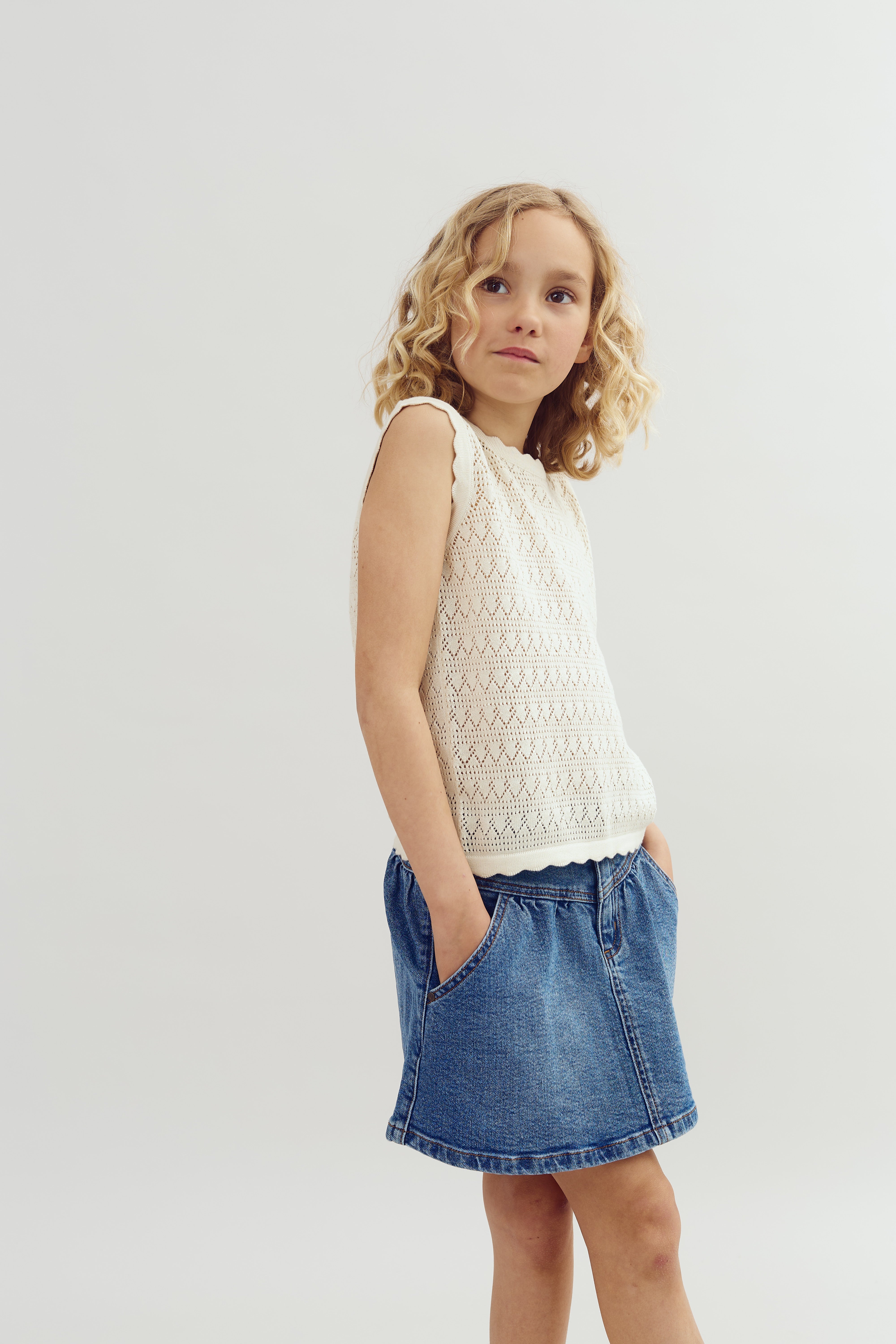 En pige med krøllet hår står med hænderne i lommerne, iført en hvid trøje og en blå denimnederdel. Baggrunden er lys og neutral.