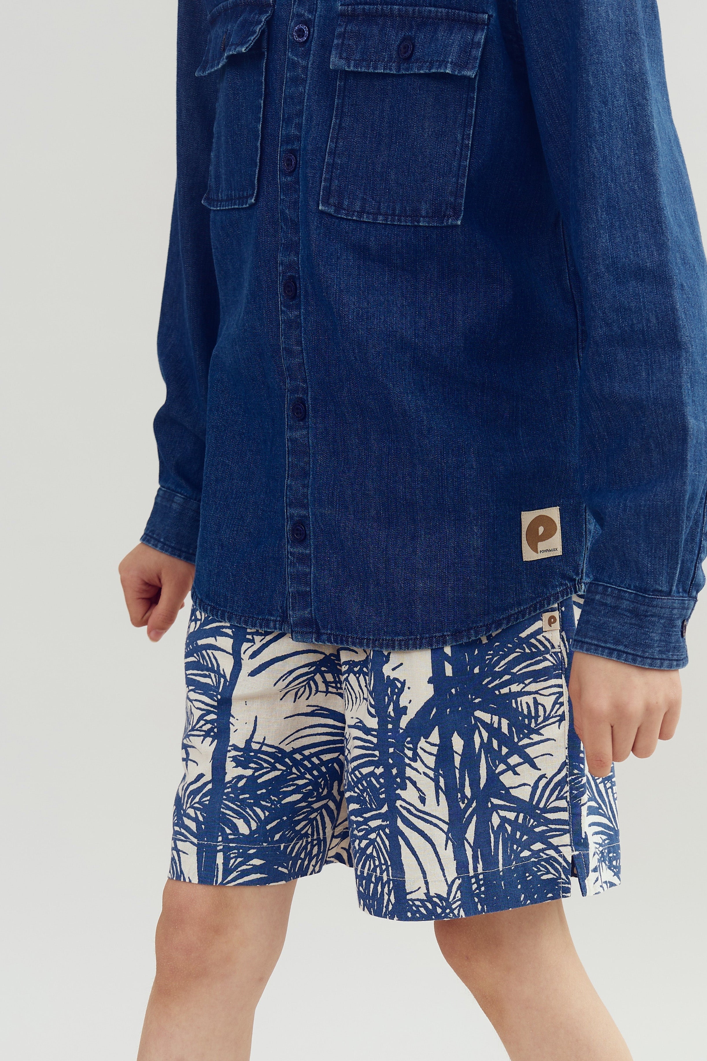 En person iført en blå denimskjorte med brystlommer og et mærke, står iført mønstrede shorts med blå bladmotiver. Baggrunden er neutral og lys.
