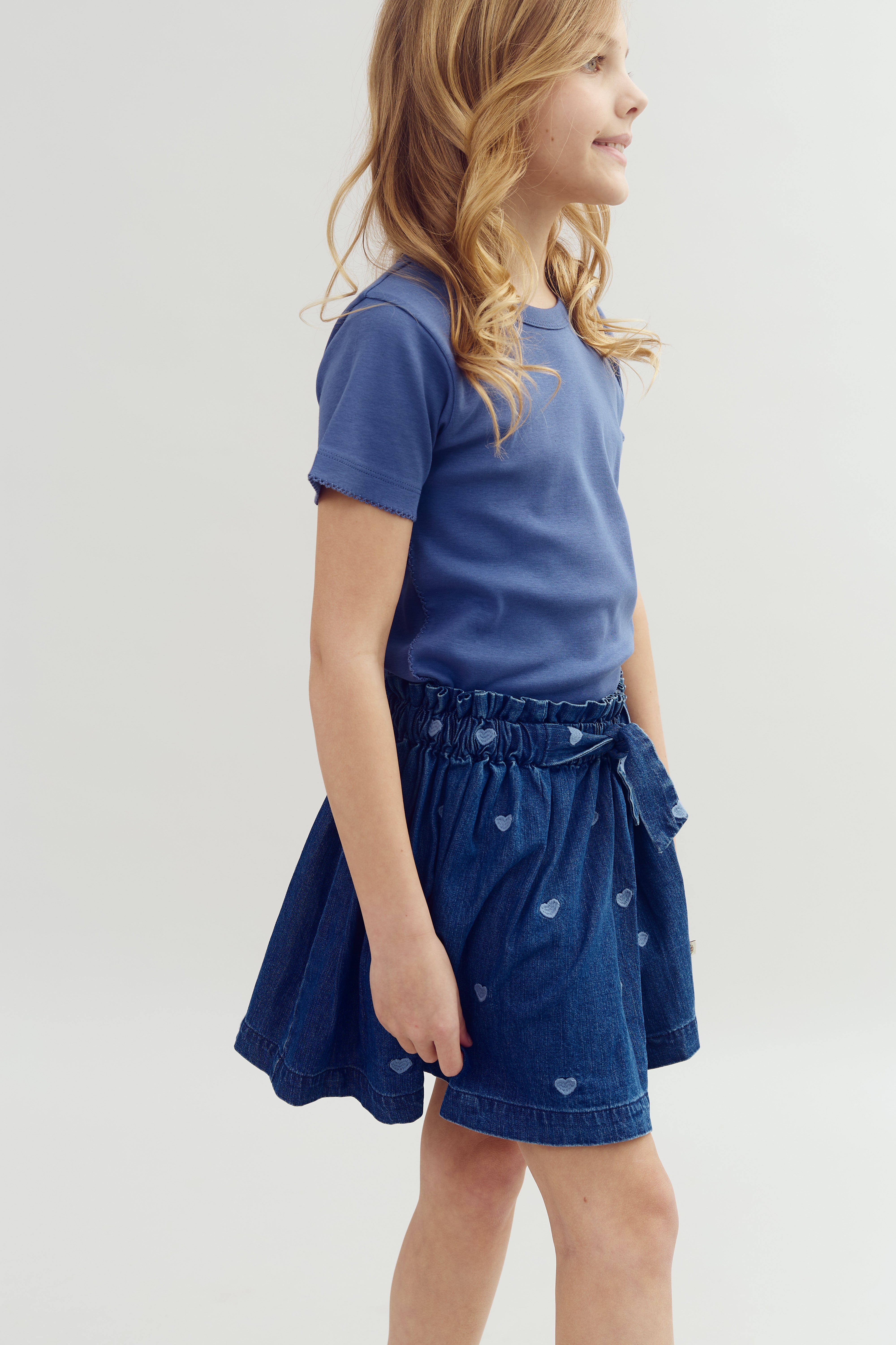 Pige står smilende, iført en blå T-shirt og en denimnederdel med hjerter. Baggrunden er neutral og lys, hvilket fremhæver hendes tøj og udtryk.