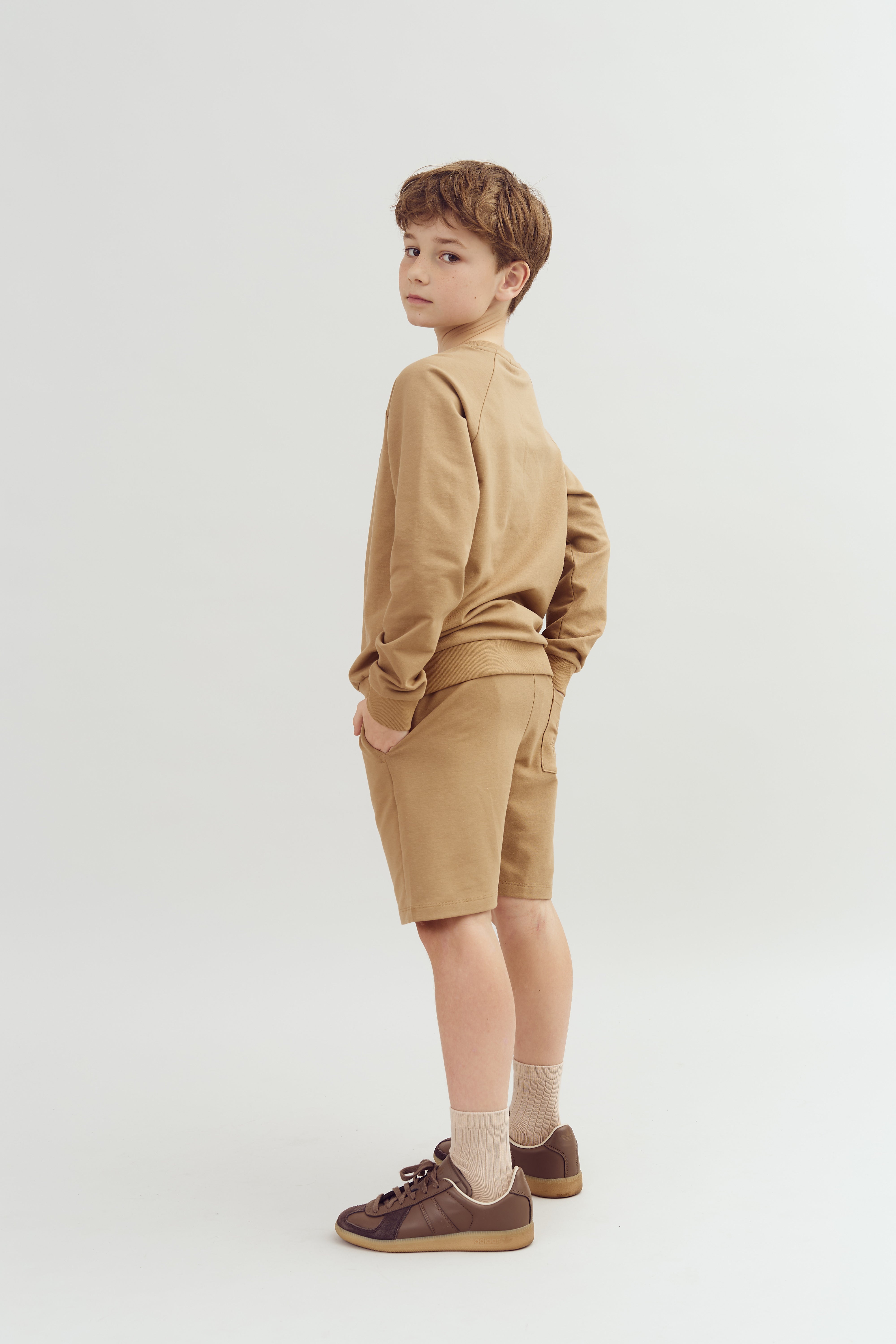 Dreng står i profil iført beige langærmet trøje og shorts, ser over skulderen i neutral, lys baggrund. Han har brune sko og sokker i matchende nuancer.