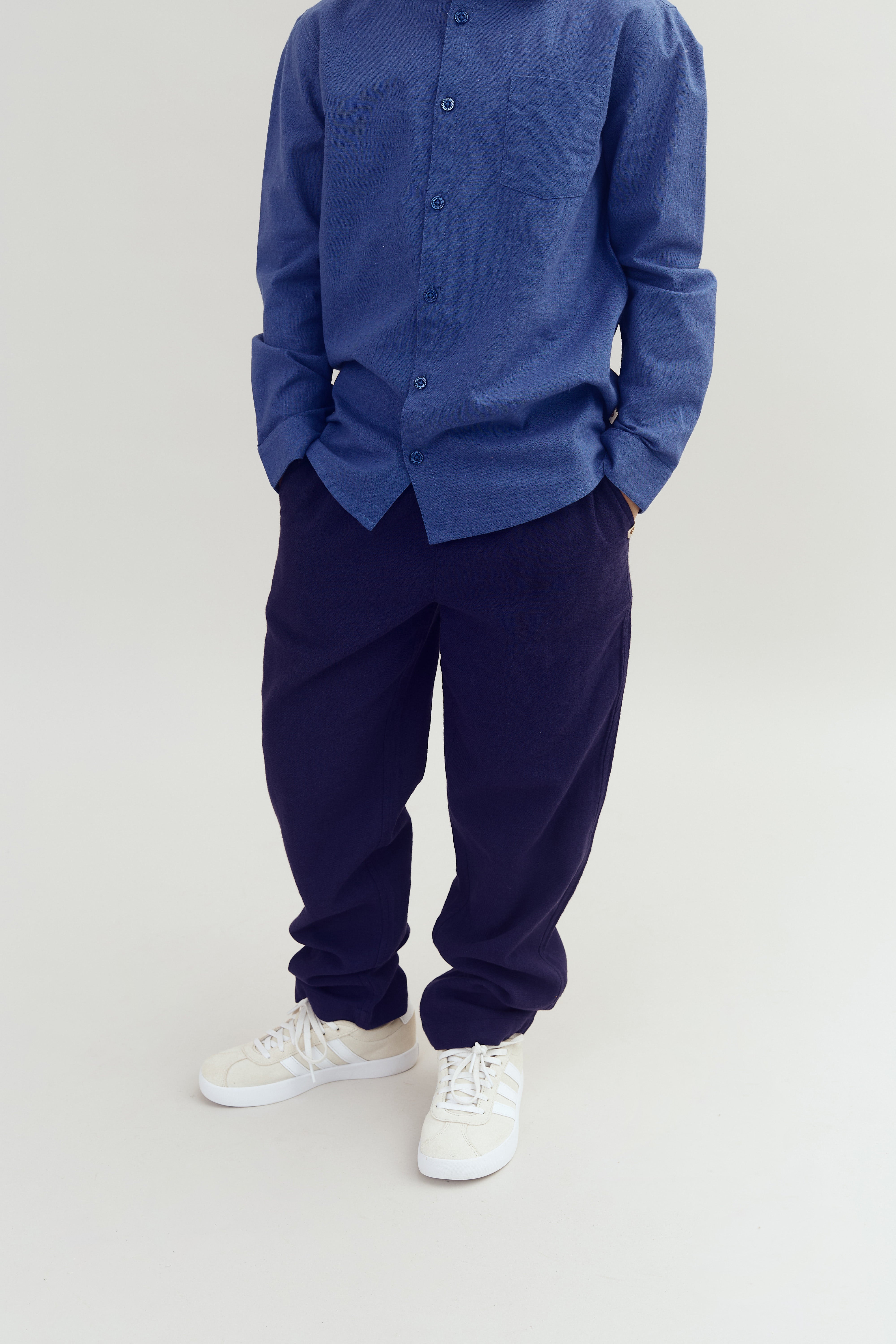 En person står med hænderne i lommerne, iført en blå skjorte, mørke bukser og hvide sneakers, mod en neutral baggrund.