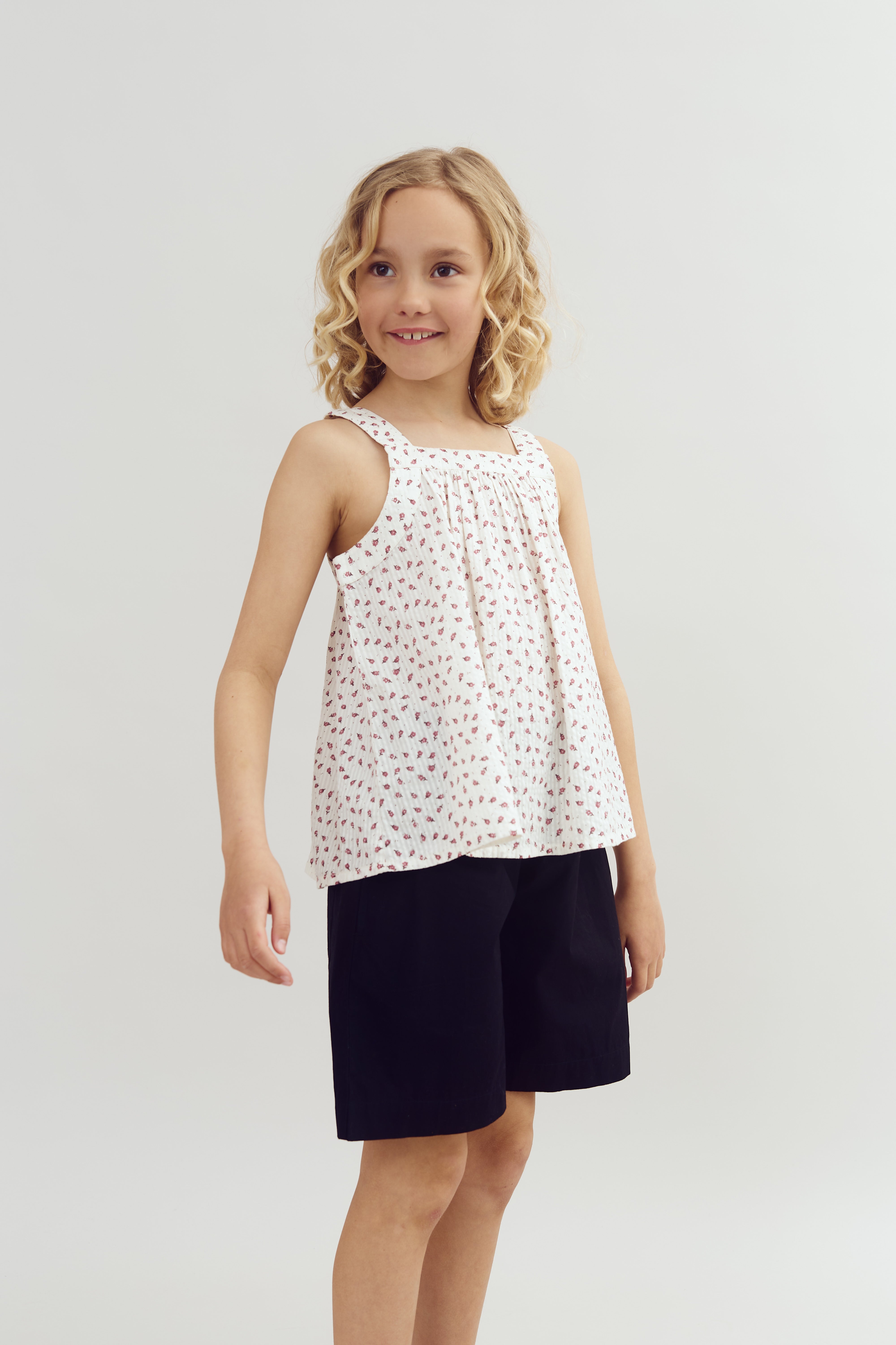 Et barn med lyst, krøllet hår står smilende iført en hvid, mønstret top og sorte shorts. Baggrunden er enkel og lys.