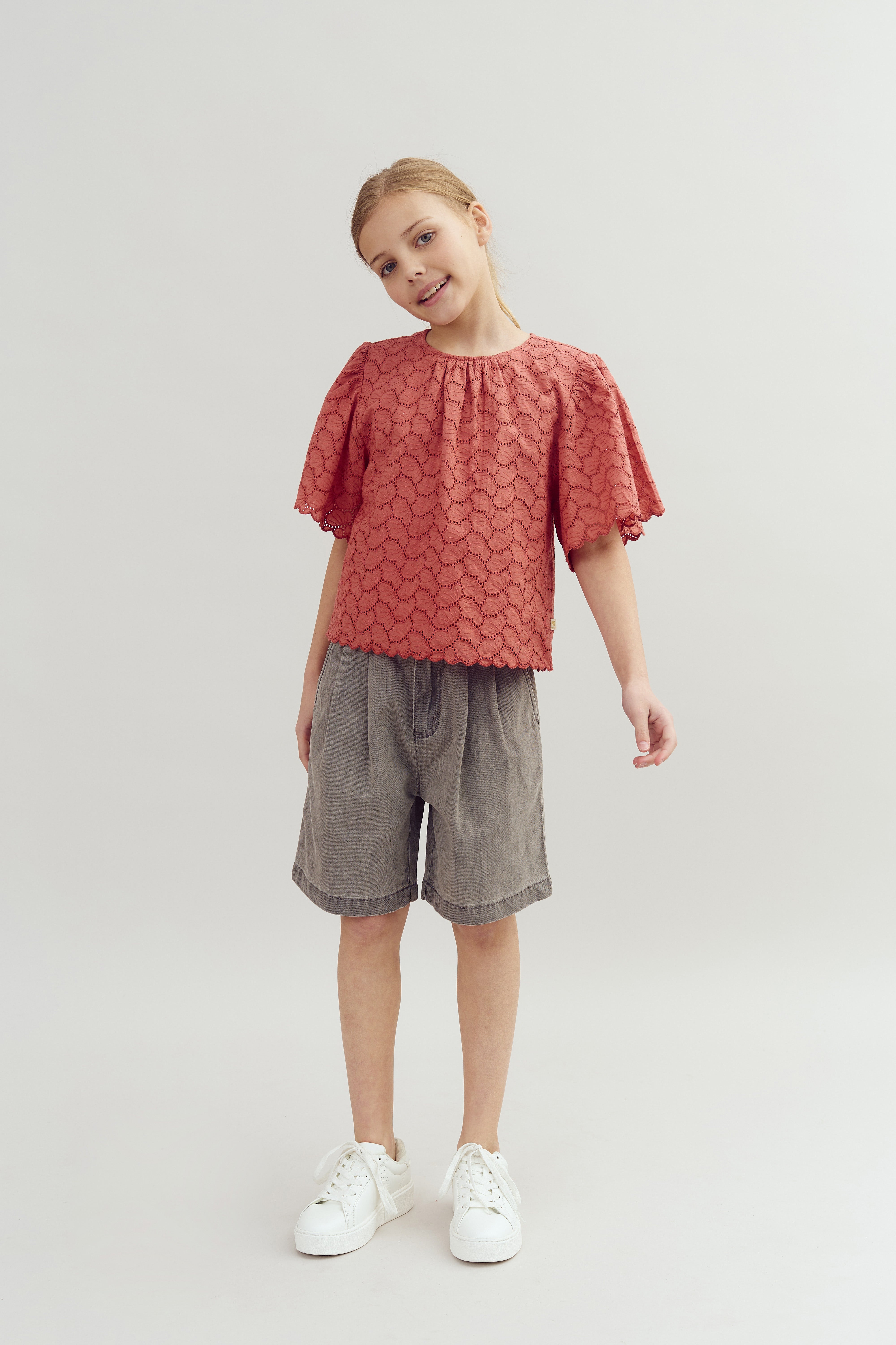 En pige står smilende iført en rød bluse med mønster og grå shorts. Hun har hvide sneakers på og står foran en neutral, lys baggrund.