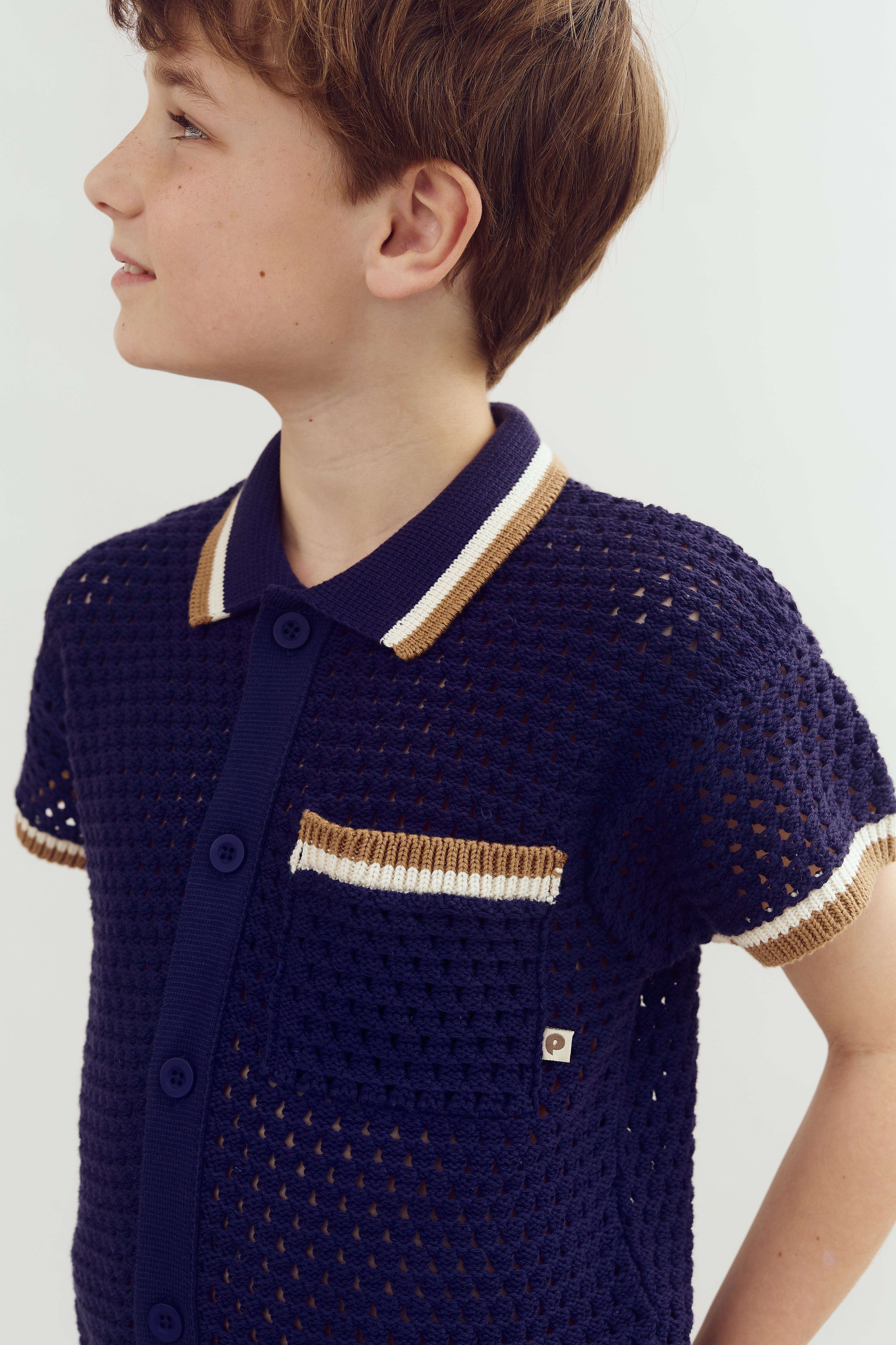 Dreng iført navyblå hæklet skjorte med brune og hvide detaljer ser til siden. Skjorten har en lomme med knapper foran. Neutral, lys baggrund.