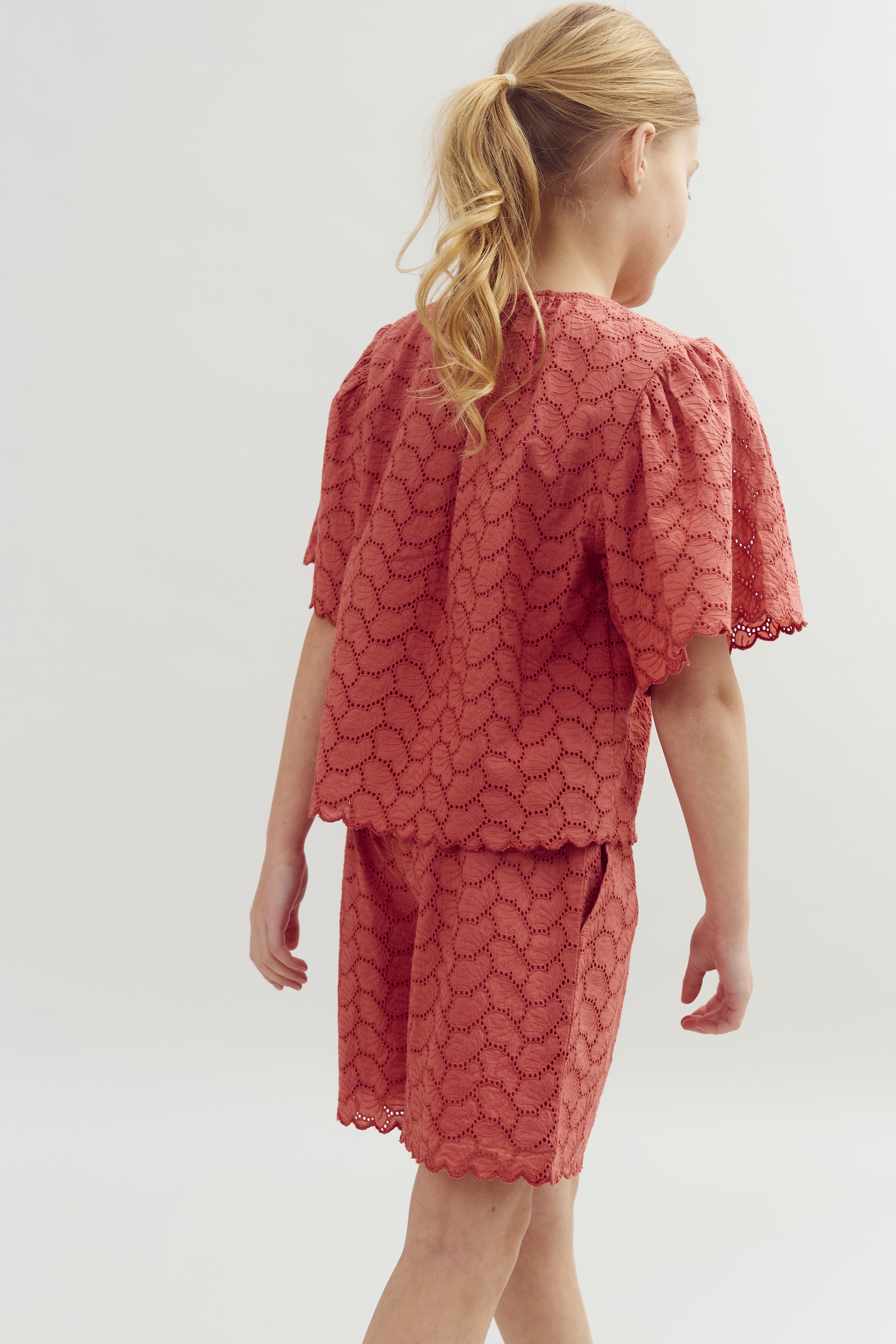 En pige iført en koralfarvet, blonderet top og shorts går væk fra kameraet i et minimalistisk, lysegråt rum. Hun har lyst hår i en hestehale.