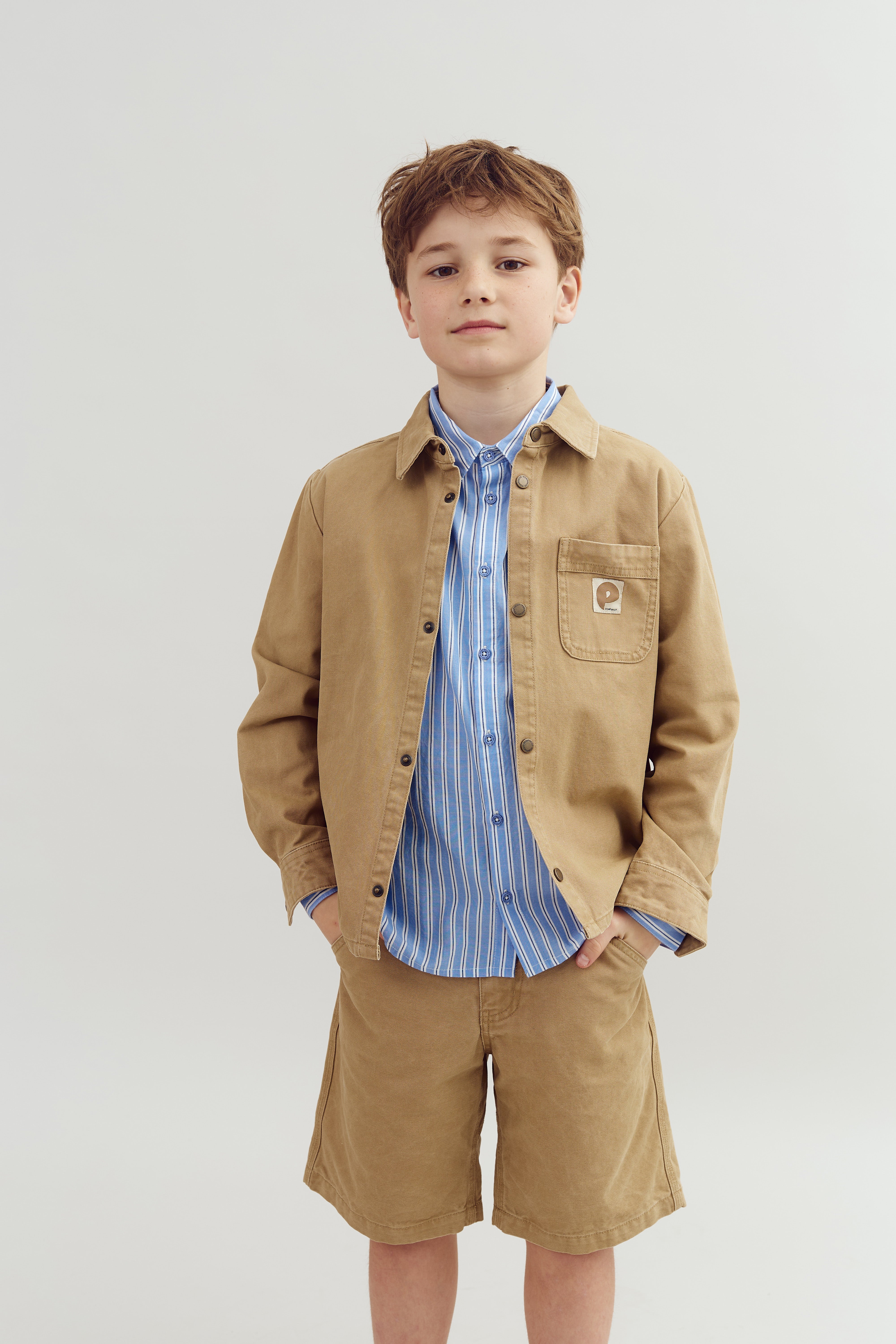 A child stands with hands in pockets, wearing a tan jacket, matching shorts, and a blue striped shirt in a neutral, plain setting. A patch on the jacket reads "Carhartt."